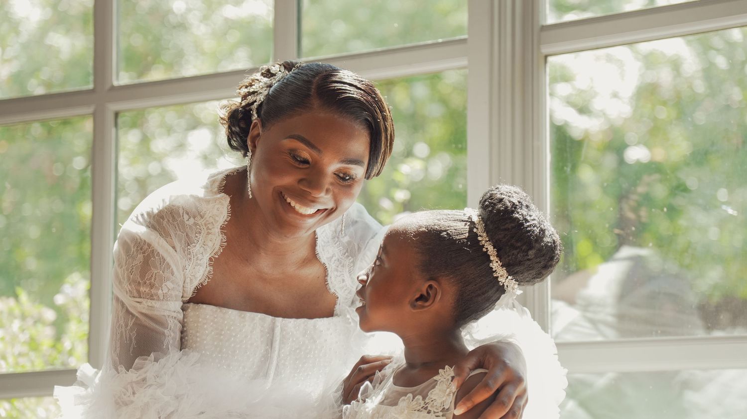 A bride in white lace smiles warmly at a young flower girl in a sunlit room.
