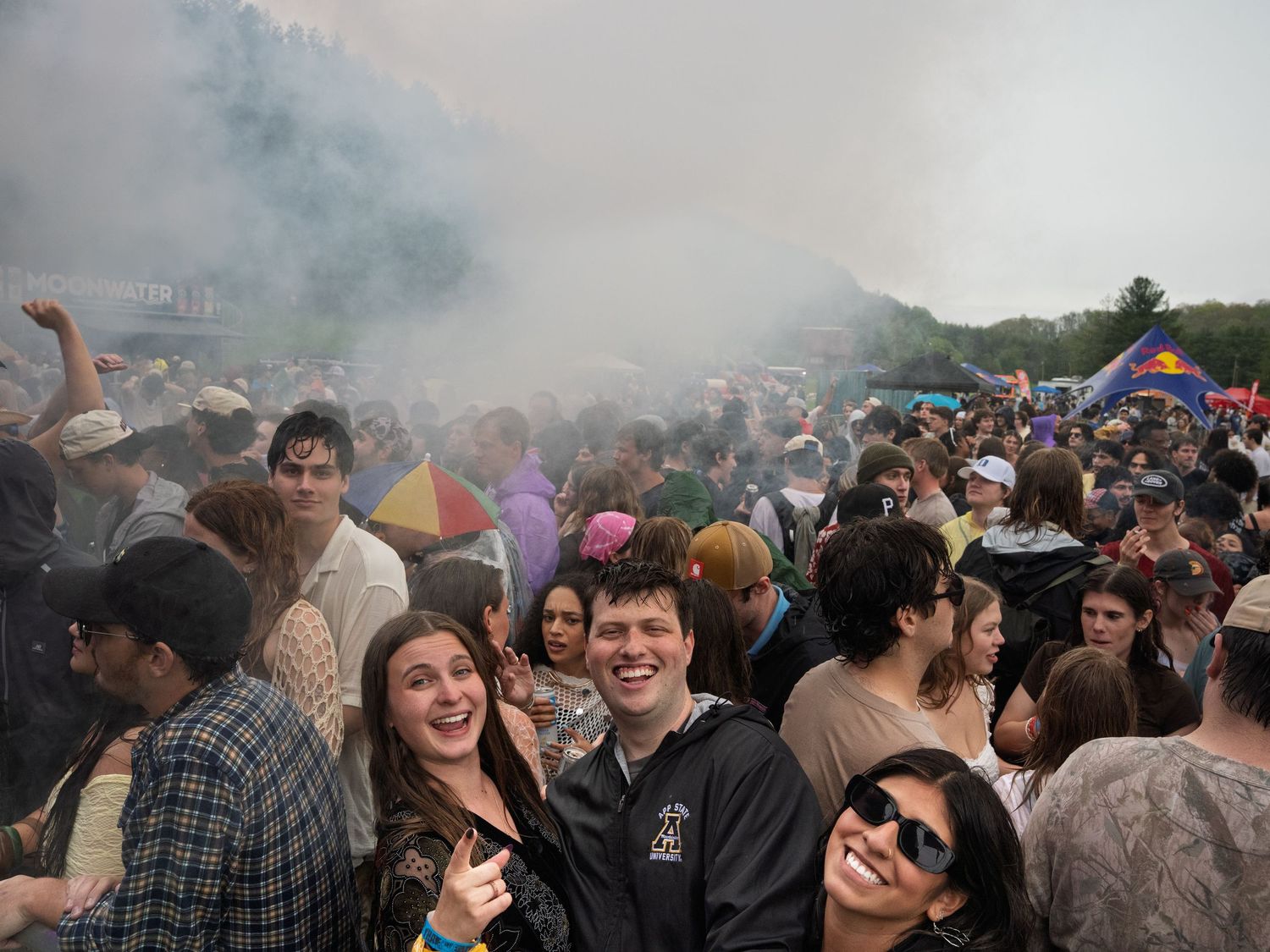 Large crowd of festival goers surrounded by smoke at outdoor music event.
