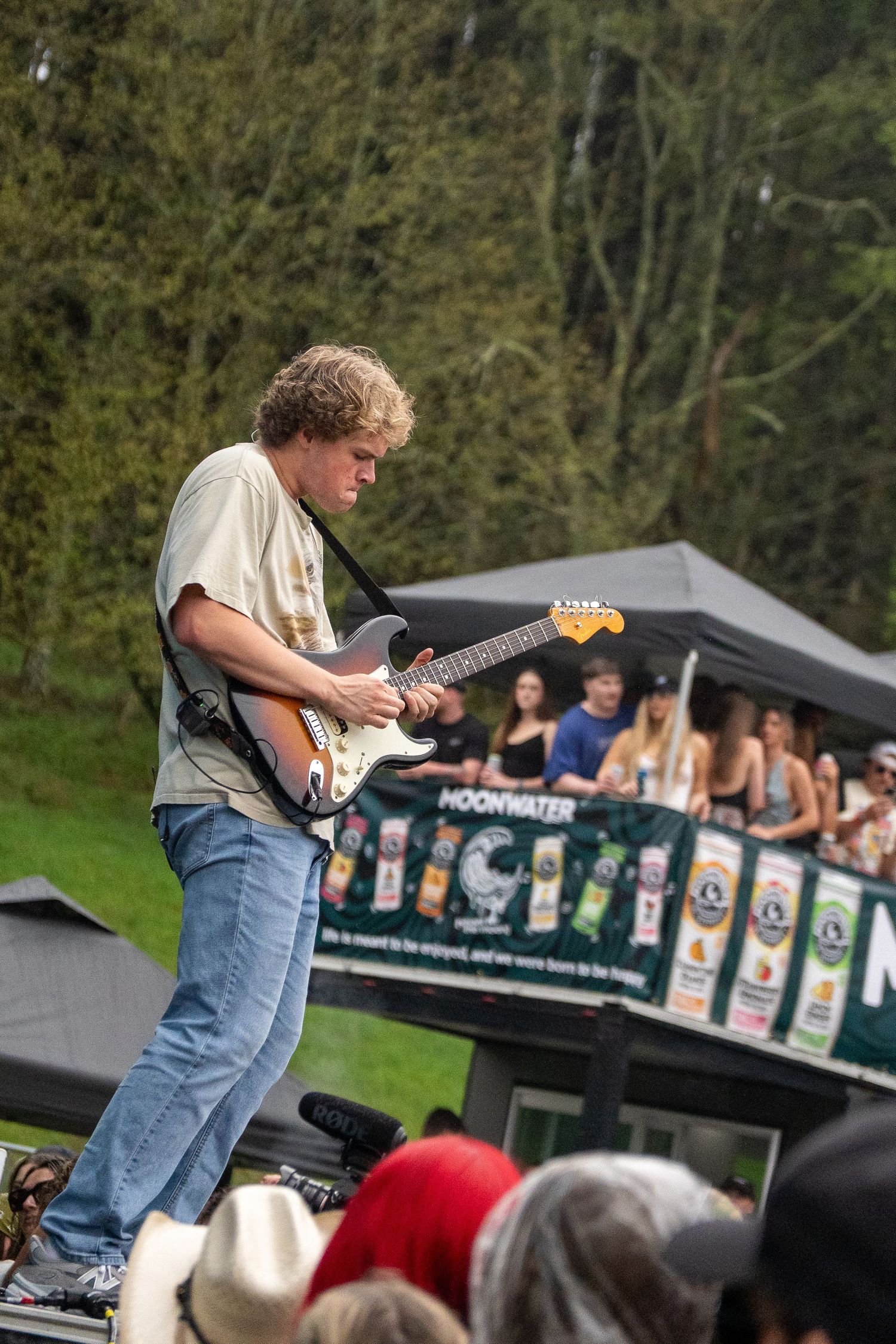 Blonde guitarist performing energetically on outdoor festival stage with crowd watching.