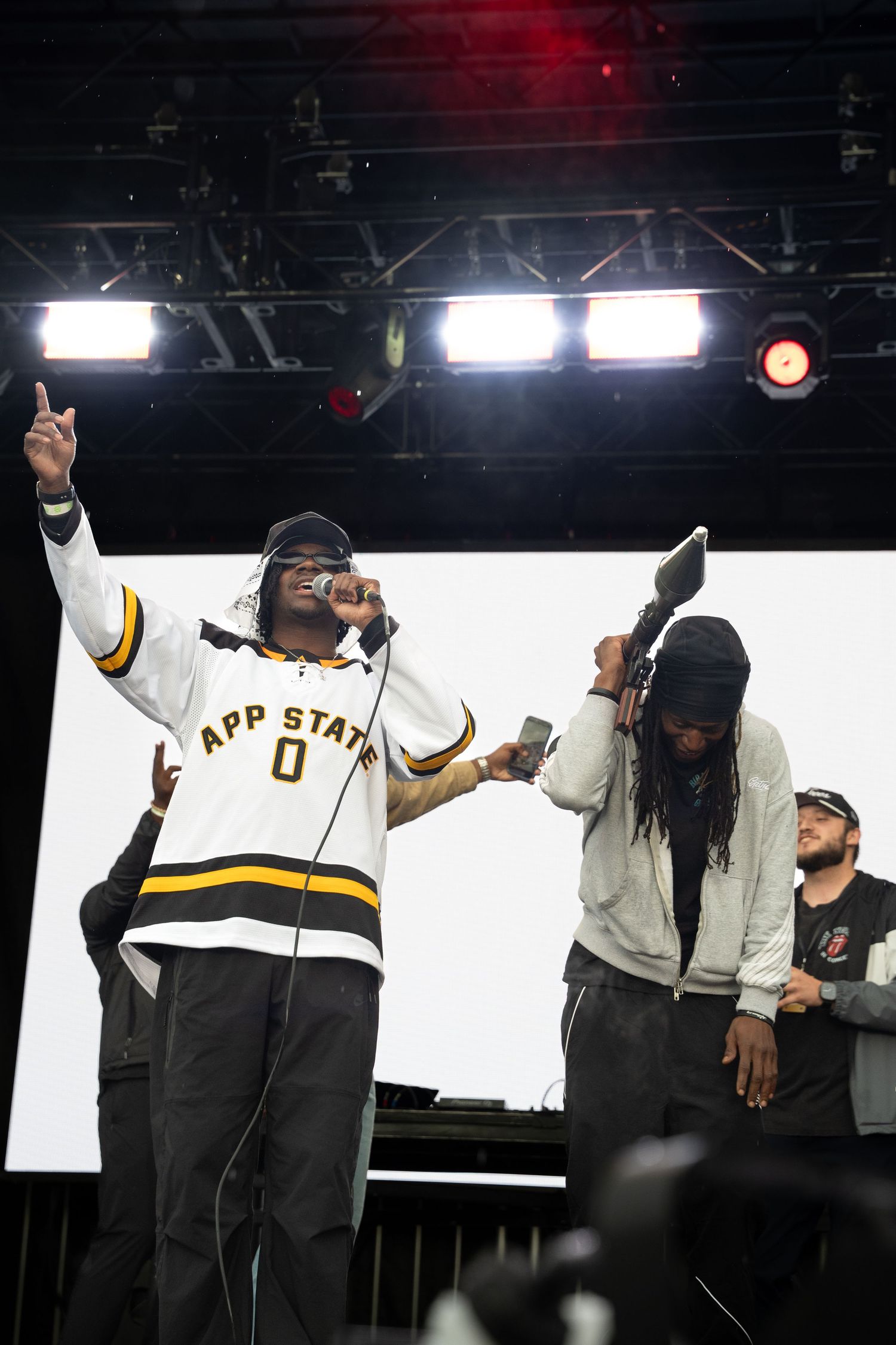 Hip-hop performer in App State jersey raises finger on smoky festival stage with crew.