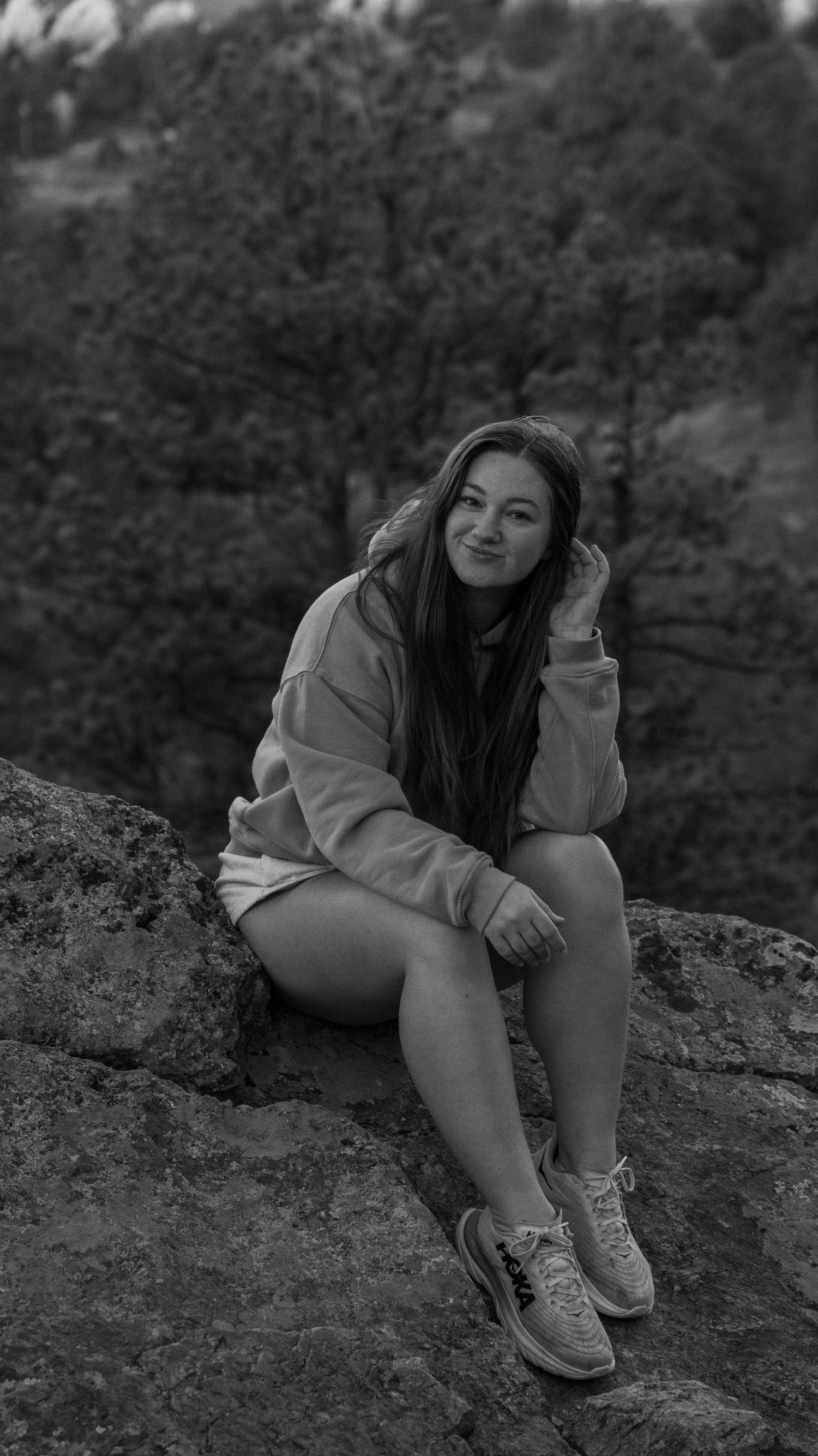 Young woman posing on rocky outcrop in black and white photo, wearing hoodie and sneakers with trees behind.
