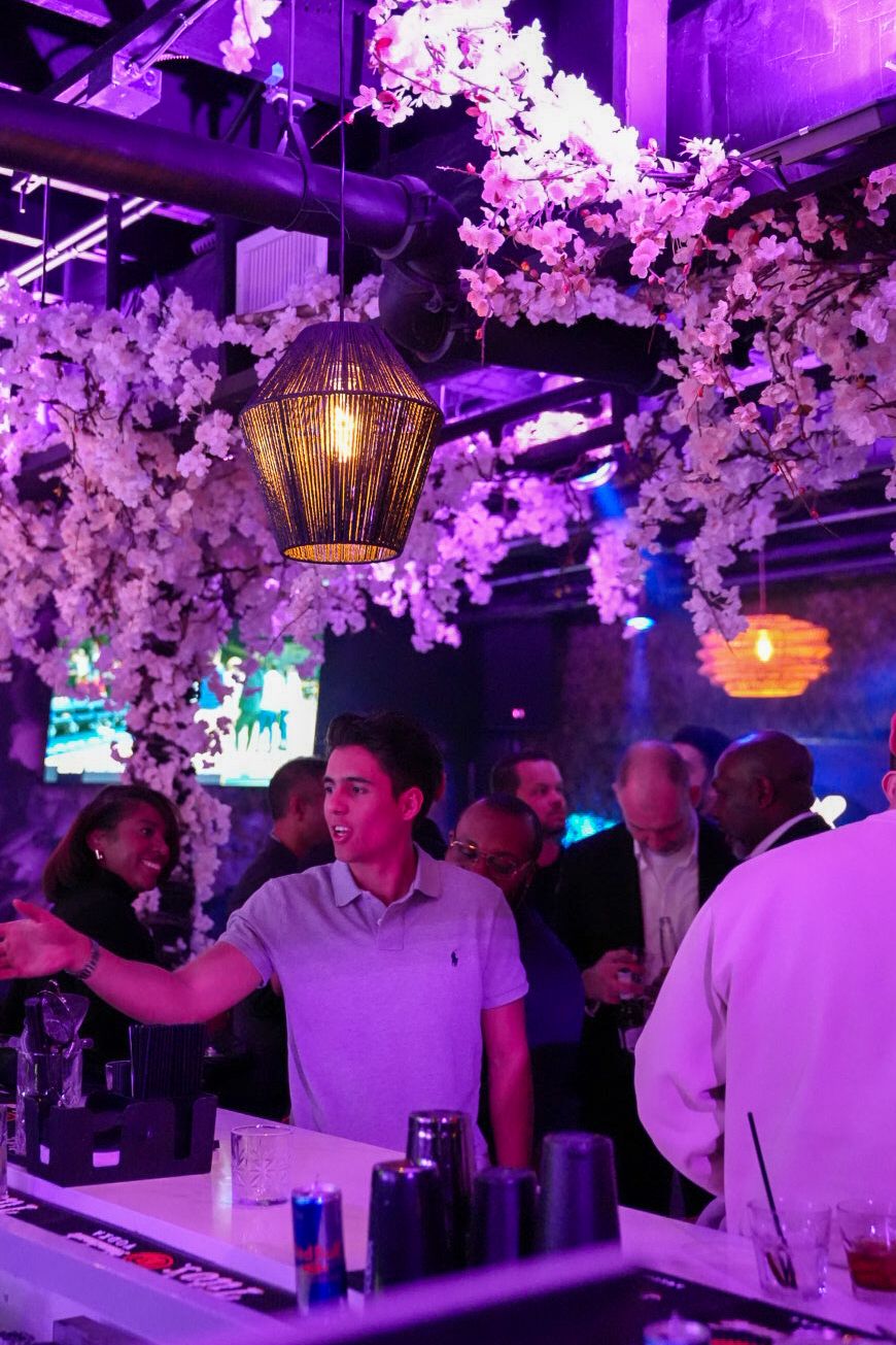 Bartender serving drinks at a bar decorated with white cherry blossom flowers under purple ambient lighting.