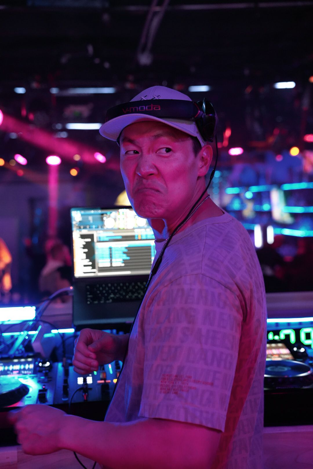 DJ wearing a cap and headphones working at a DJ booth under colorful purple and pink neon club lights.