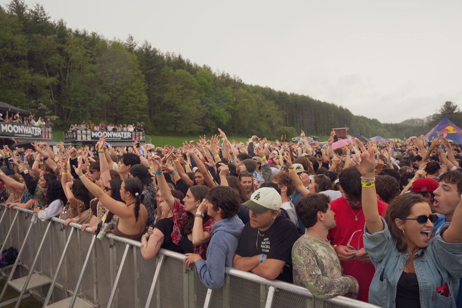 Large crowd of festival-goers with hands raised at an outdoor music festival near a forested hillside.