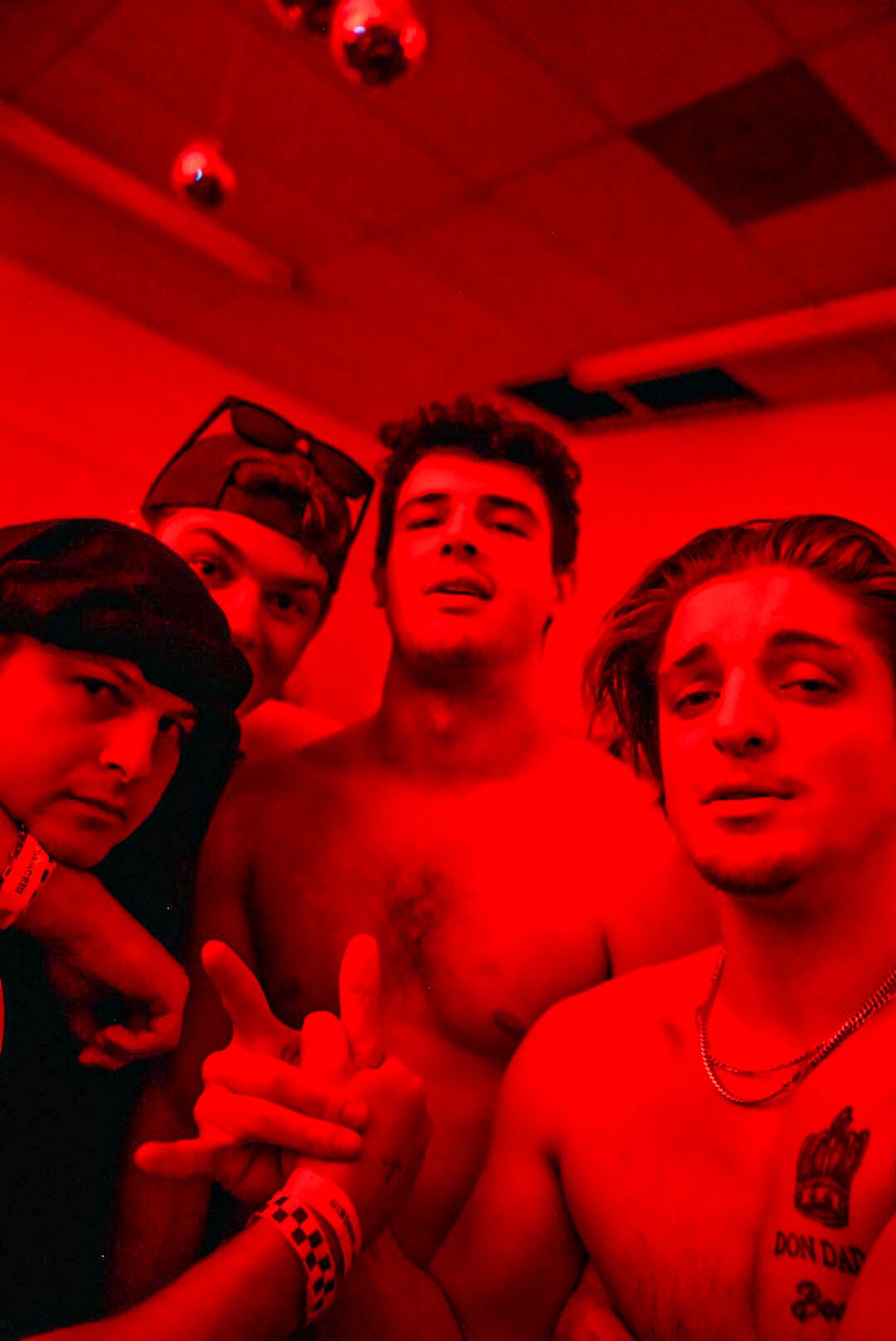 Four shirtless young men pose together under intense red lighting at a nightclub party.