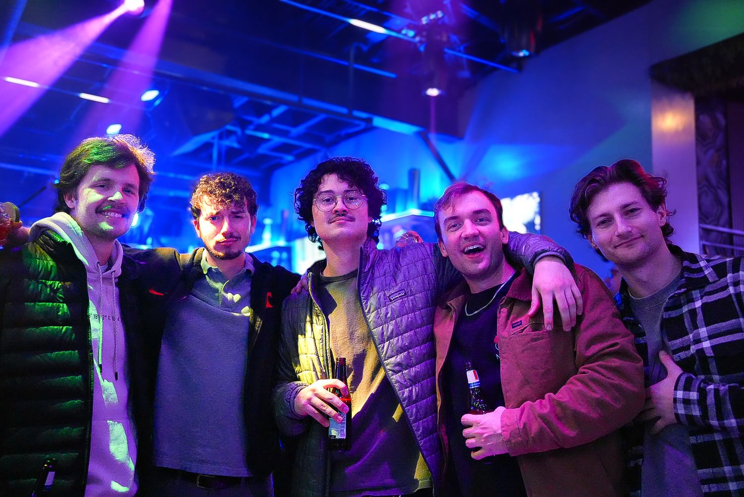 Group of five young men smiling together at a nightclub with blue stage lighting.