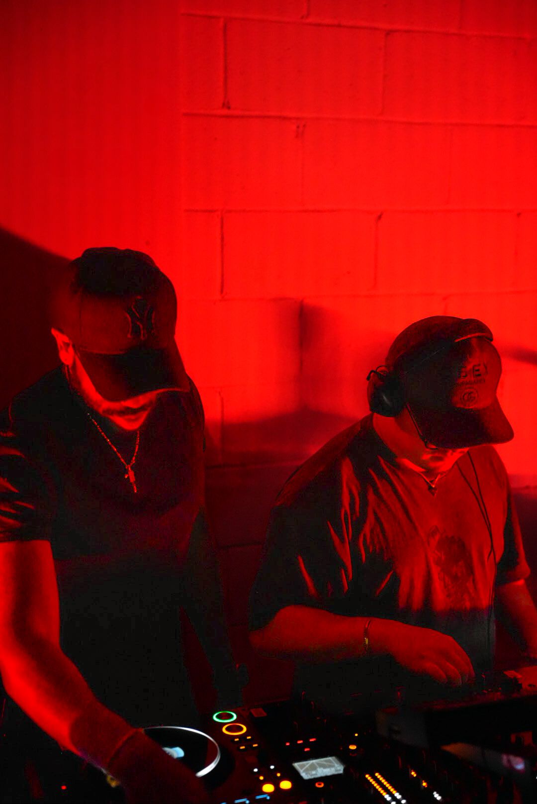 Two DJs wearing caps work Pioneer DJ equipment under dramatic red lighting at a nightclub event.