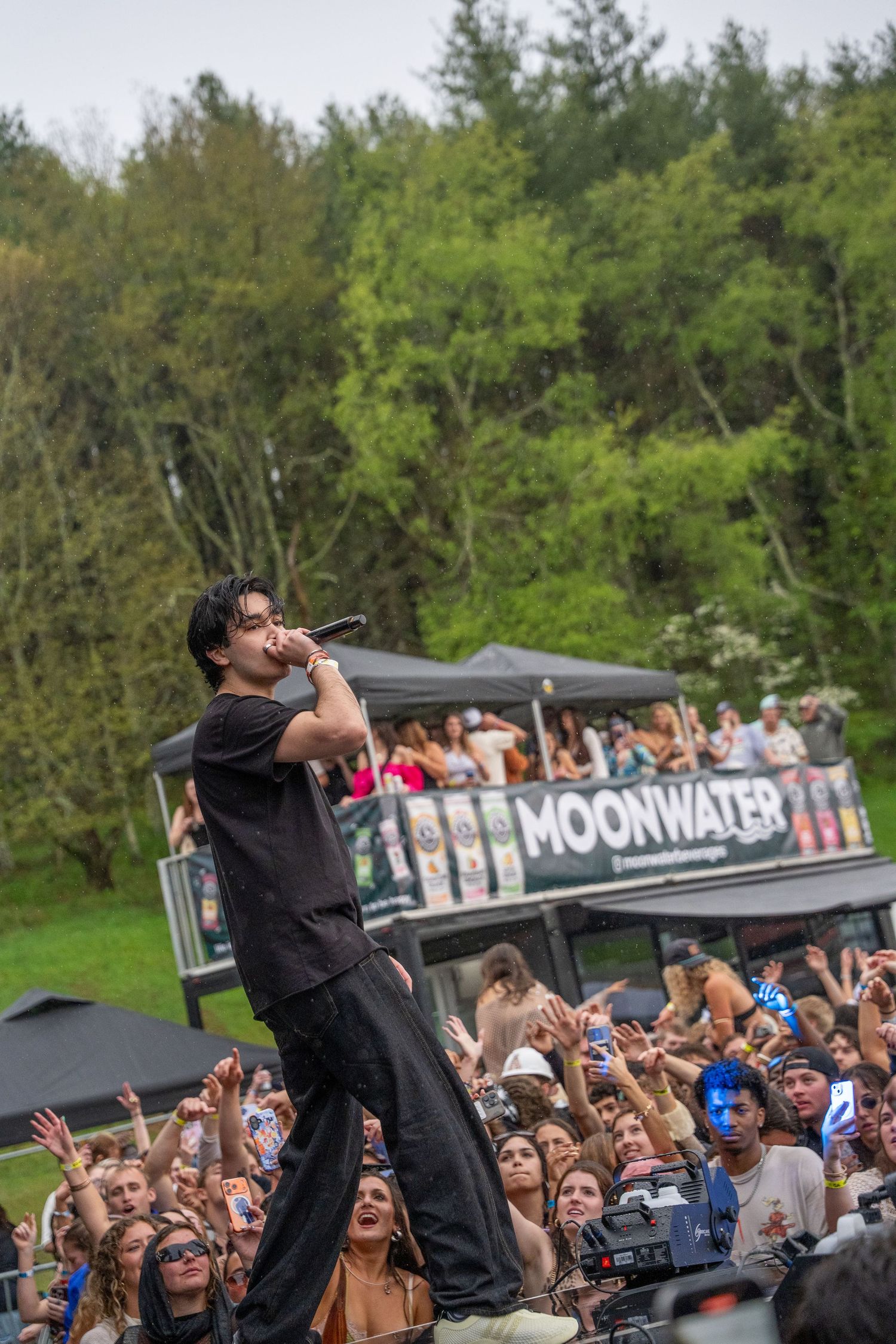 A male artist sings into a microphone on stage before a large crowd at a Moonwater outdoor festival.