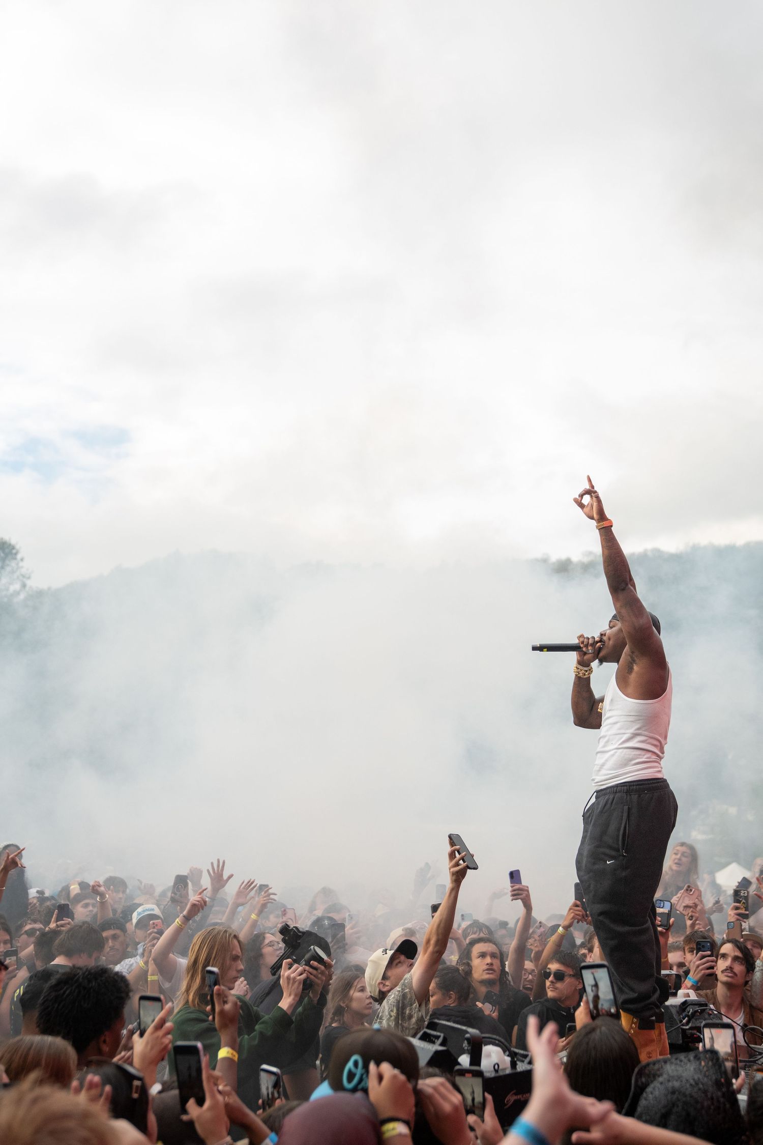 Rapper in white tank top points skyward amid smoke at packed outdoor music festival.