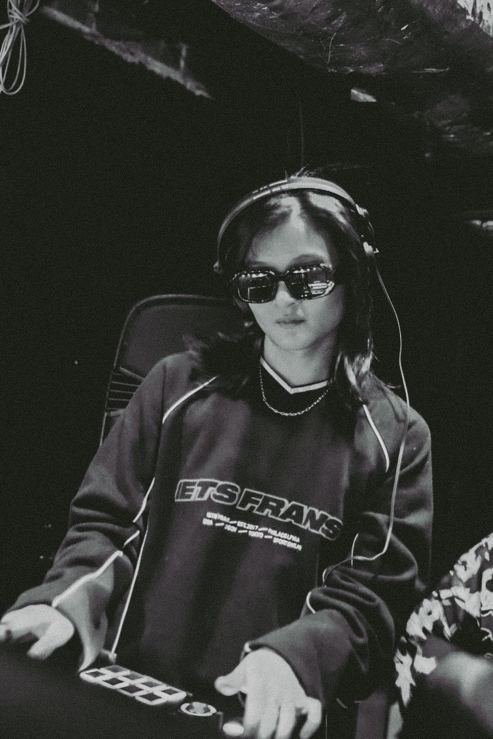 Black and white photo of a female DJ wearing sunglasses and headphones mixing music at a nightclub.
