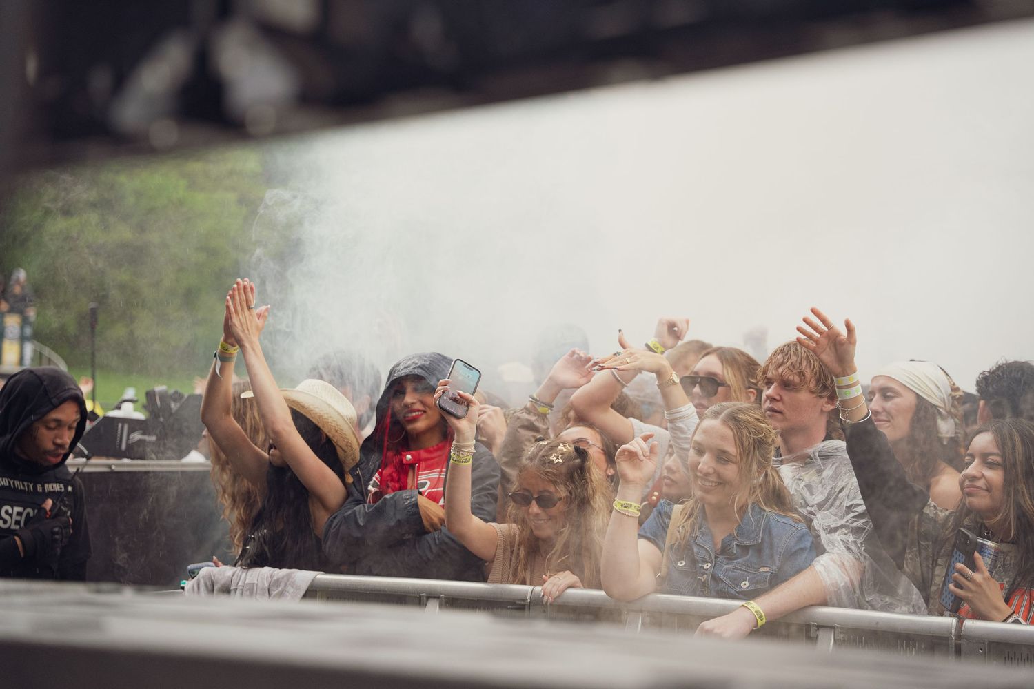 Crowd of excited concert-goers with raised hands near a smoky outdoor festival stage.