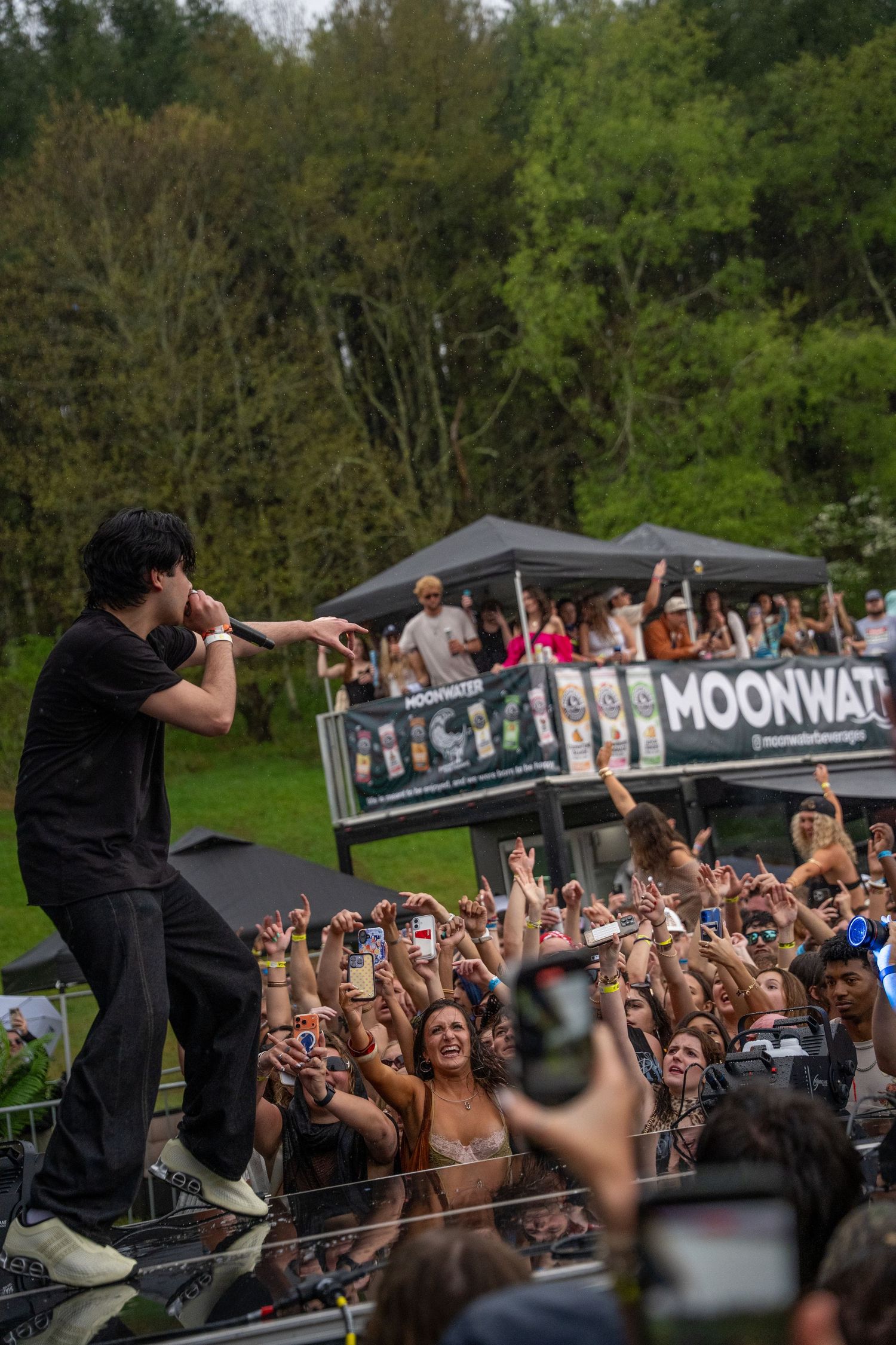 A performer on stage points to an energetic crowd at a Moonwater sponsored outdoor festival.