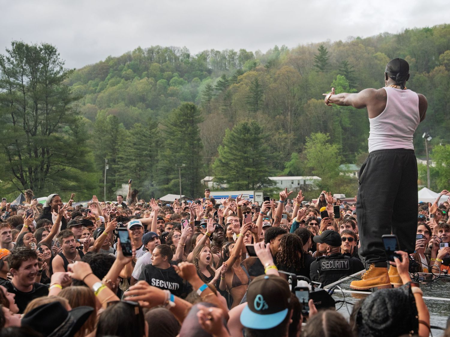 Performer in white tank top gestures to massive crowd at outdoor music festival.