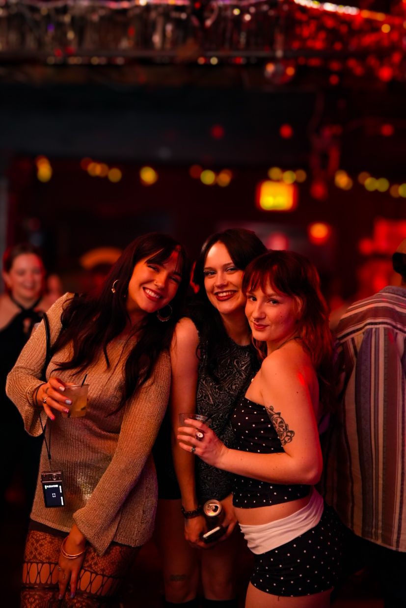 Three women smiling and posing together at a dimly lit nightclub with colorful bokeh lights in background.