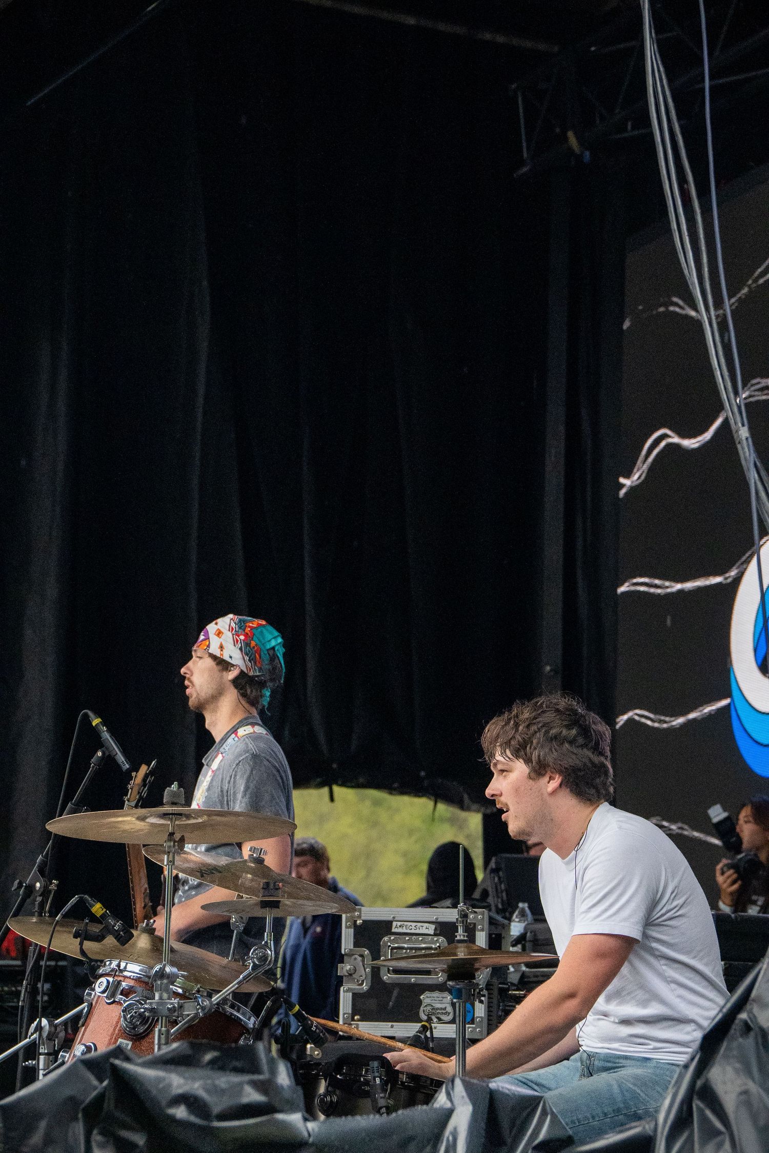 Two musicians performing on outdoor festival stage, one playing drums with bandana headwrap.