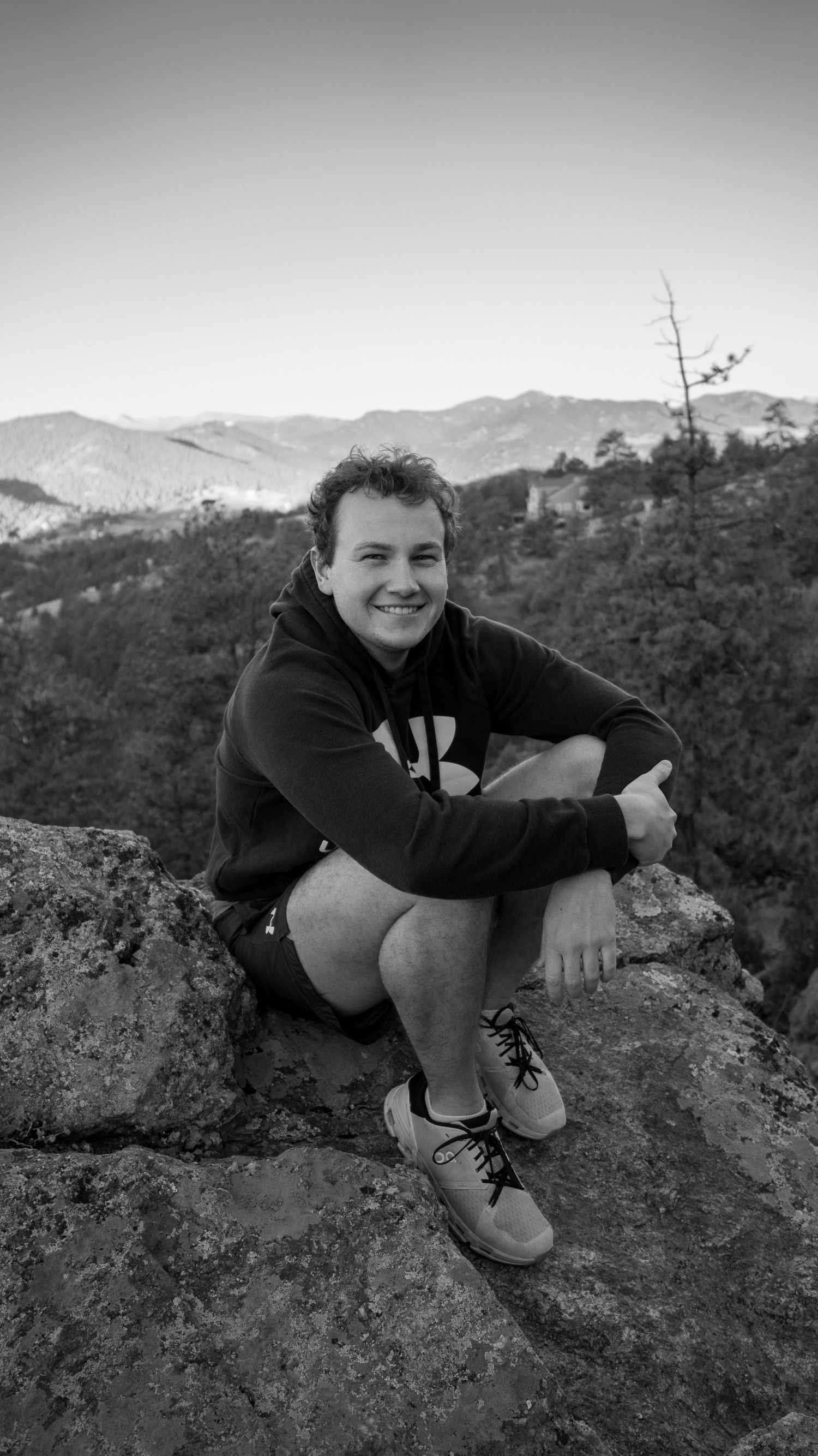 Young man sitting on rocky mountain overlook, smiling in black and white portrait with forest backdrop.