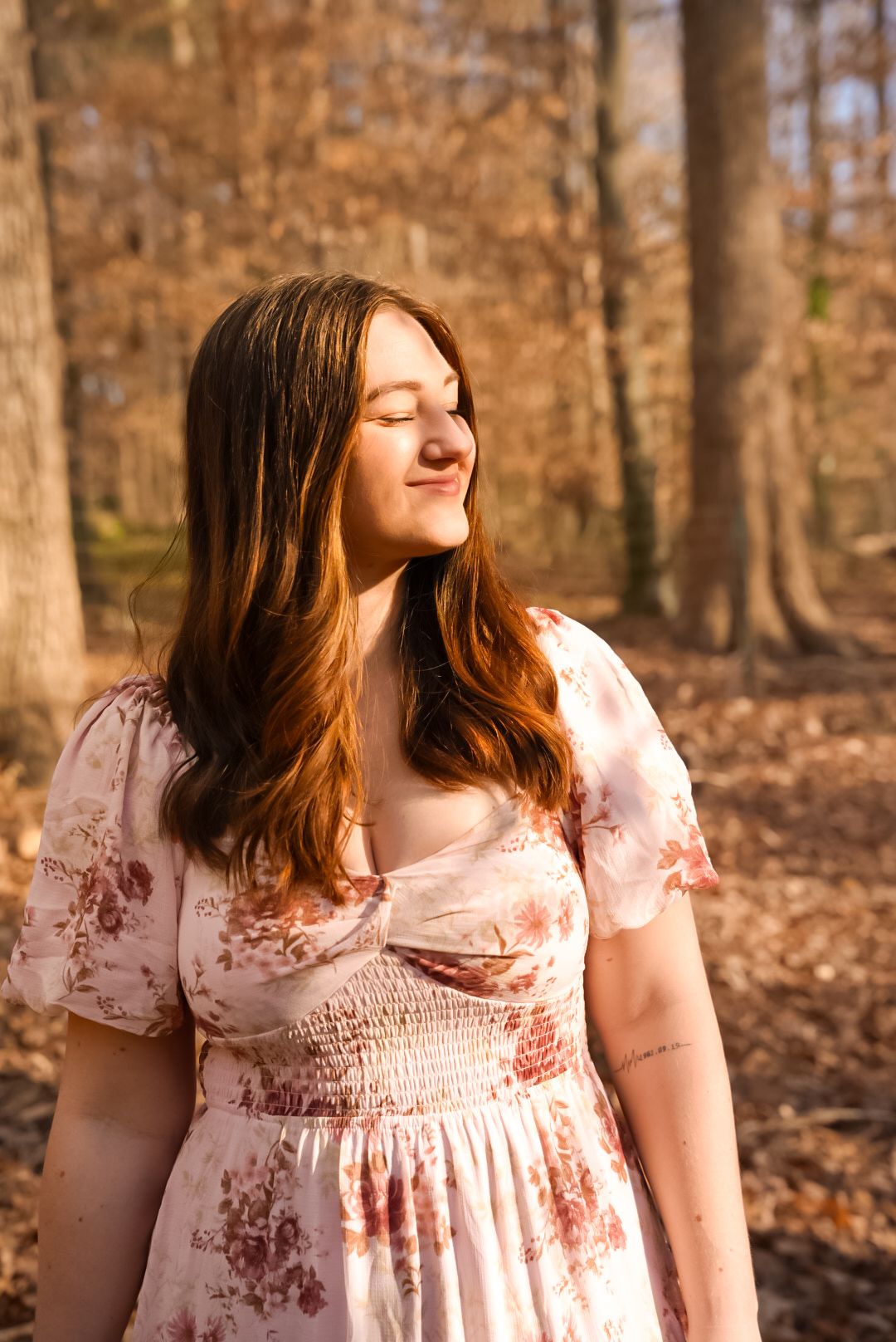 Young woman in floral dress standing peacefully in autumn forest with golden sunlight.