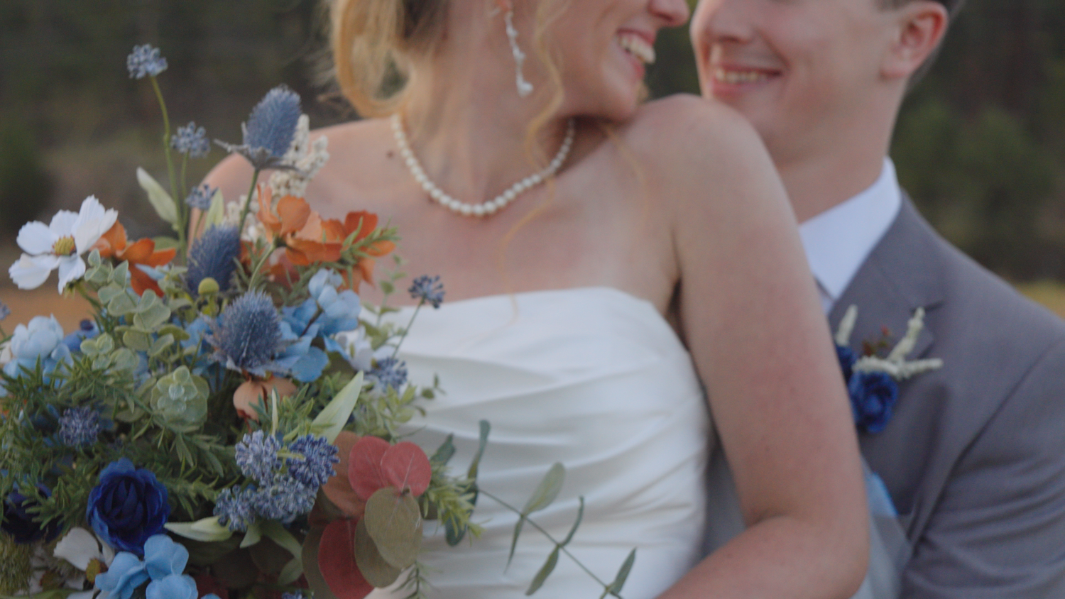 Close-up of bride and groom embracing with colorful blue and orange wedding bouquet outdoors.