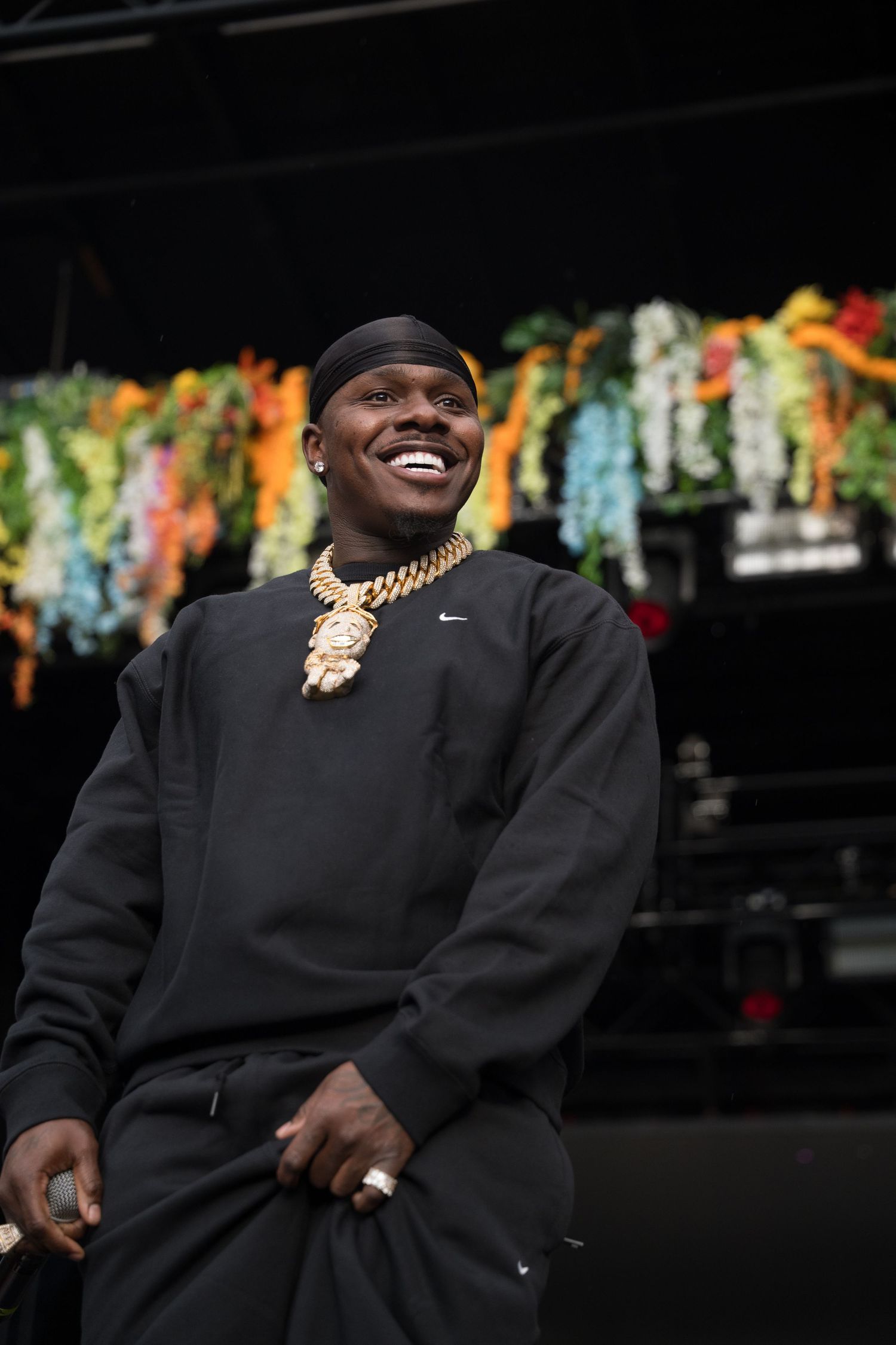 Cheerful rapper wearing black Nike sweatshirt and large gold chain smiles broadly in front of colorful festival stage decor.