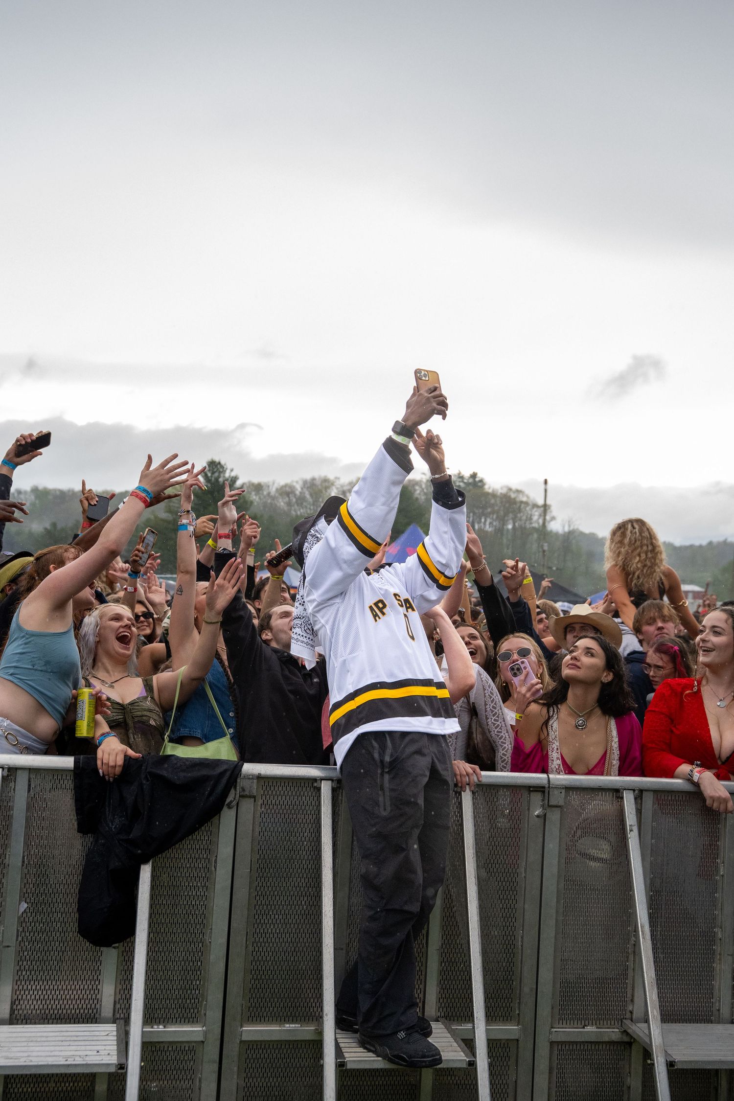 Person in white hockey jersey stands on barrier taking selfie with reaching festival crowd.