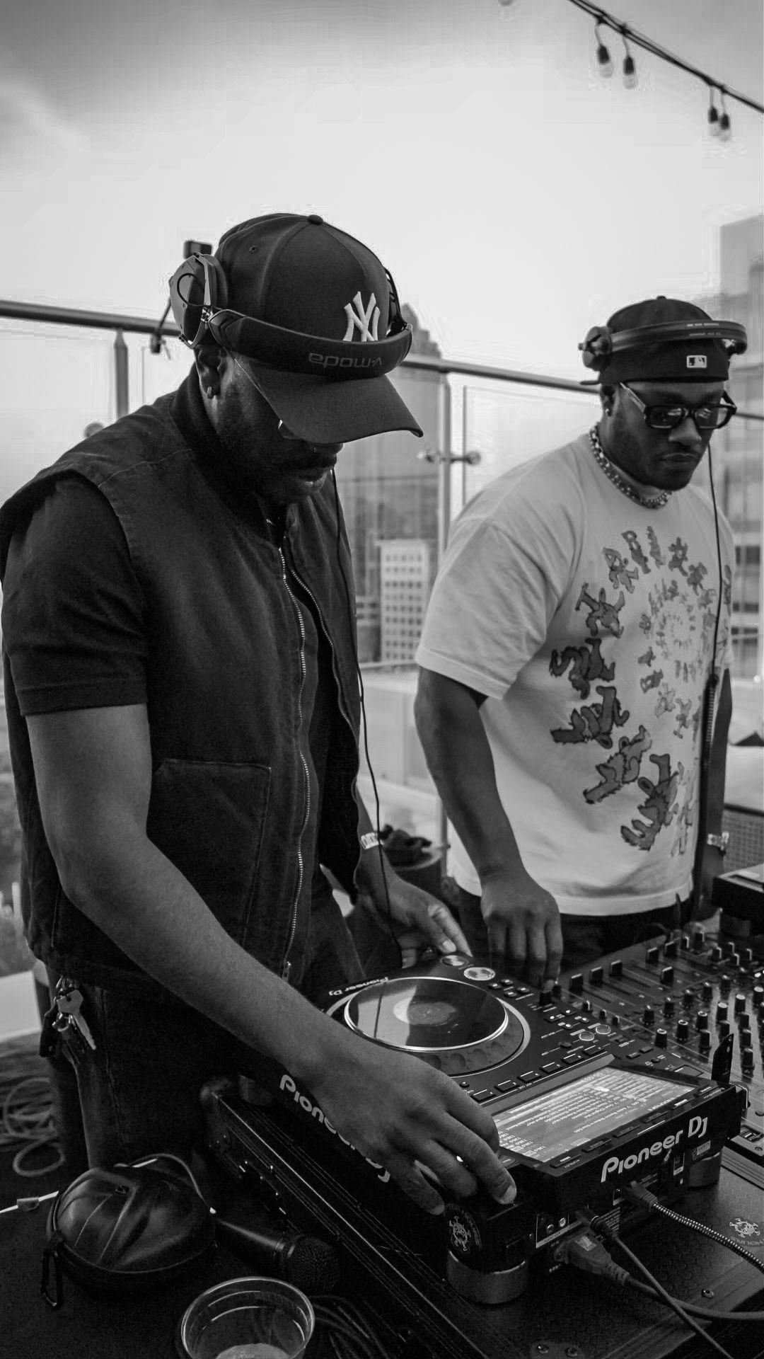 Two DJs performing on rooftop venue using Pioneer DJ equipment in black and white urban setting.