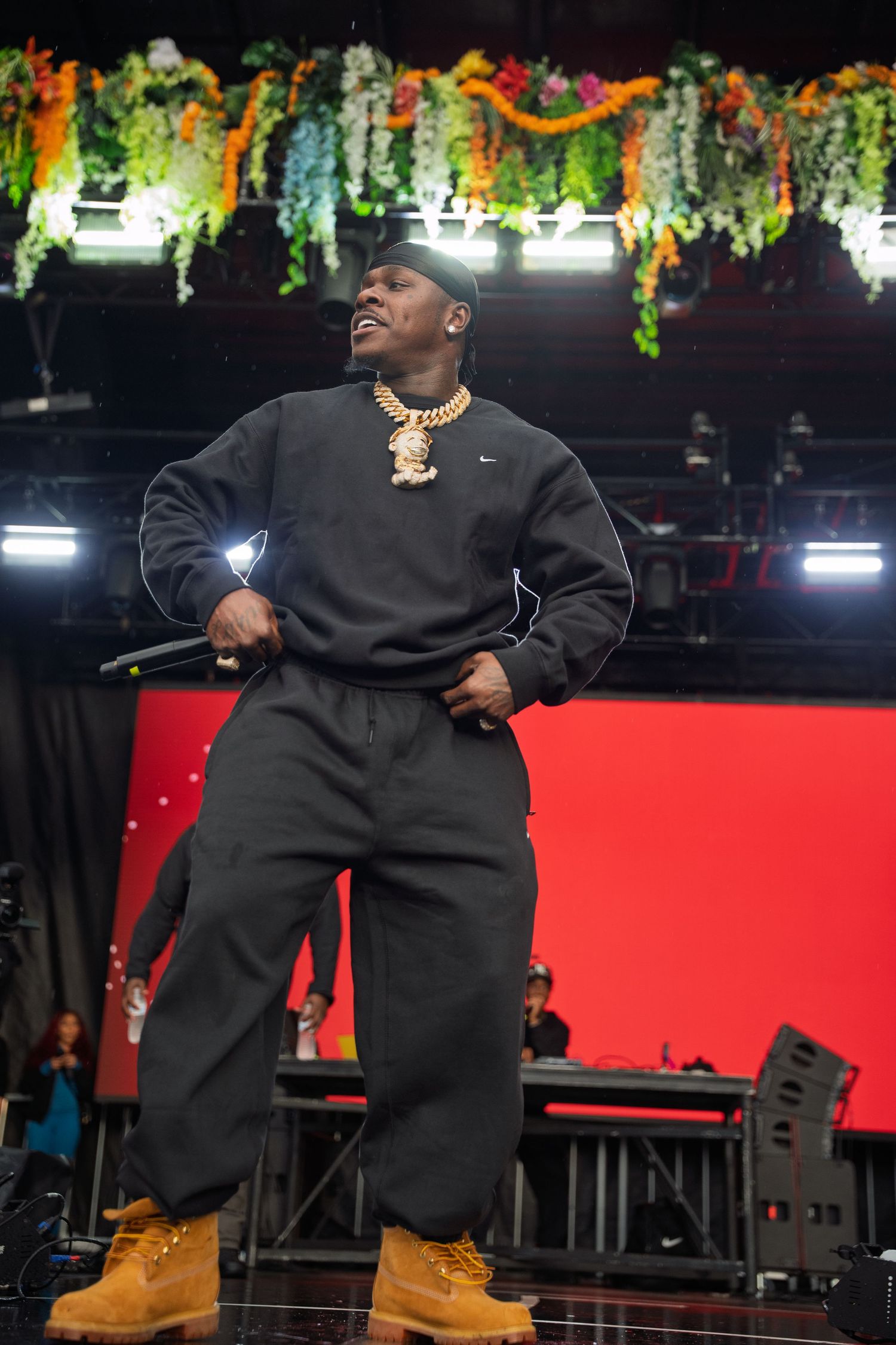 Rapper in black tracksuit and Timberland boots performs energetically on stage decorated with colorful hanging floral garlands.