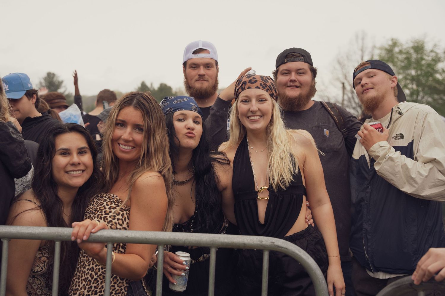 Group of smiling friends posing together at an outdoor music festival near a metal barrier.