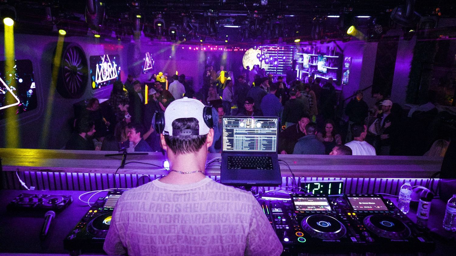 DJ performing on stage with laptop and turntables before a large purple-lit crowd.