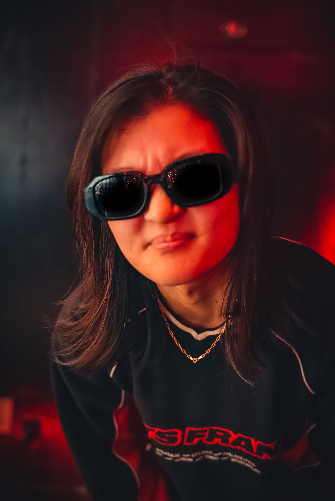 Young woman in dark sunglasses wearing black streetwear under red lighting.