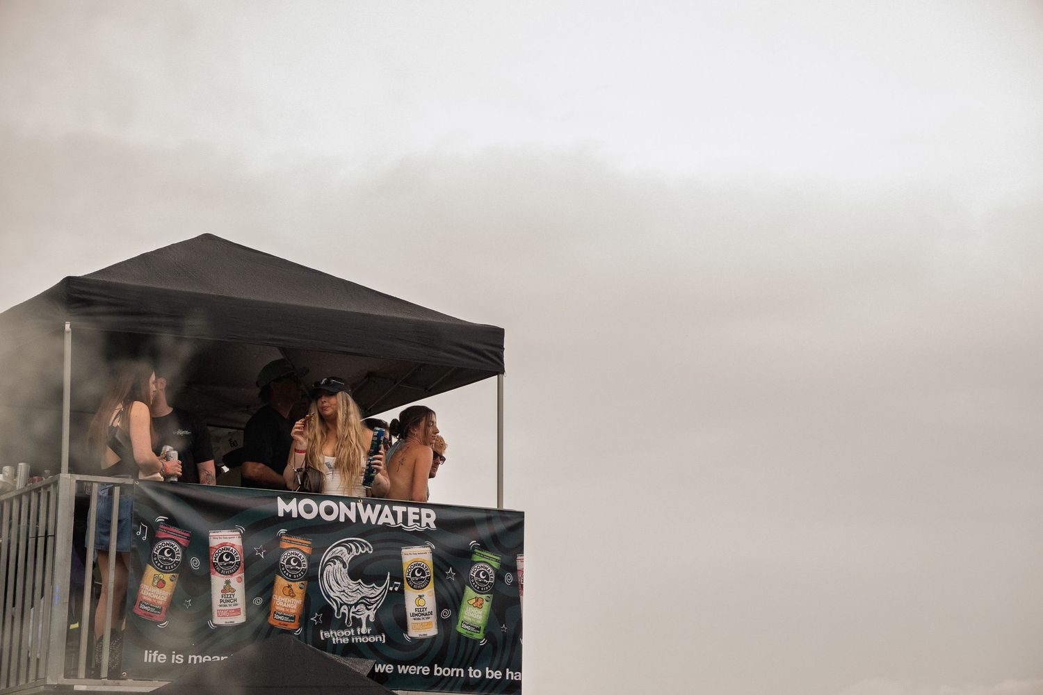 People standing on Moonwater sponsored elevated platform at foggy outdoor music festival.