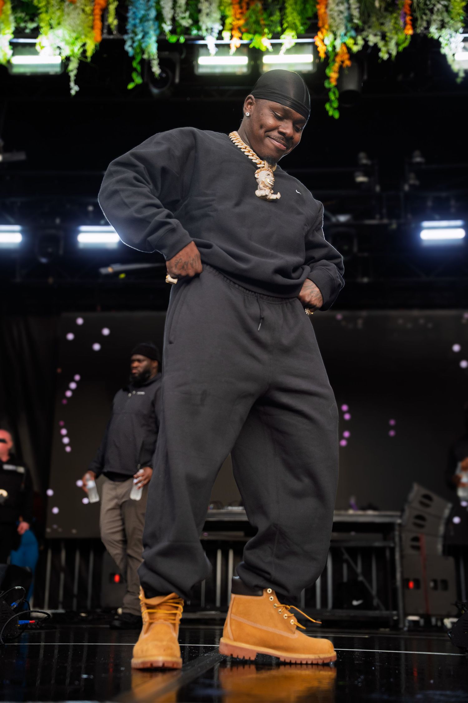 Rapper in black tracksuit and tan Timberland boots strikes a confident pose on colorfully decorated festival stage.