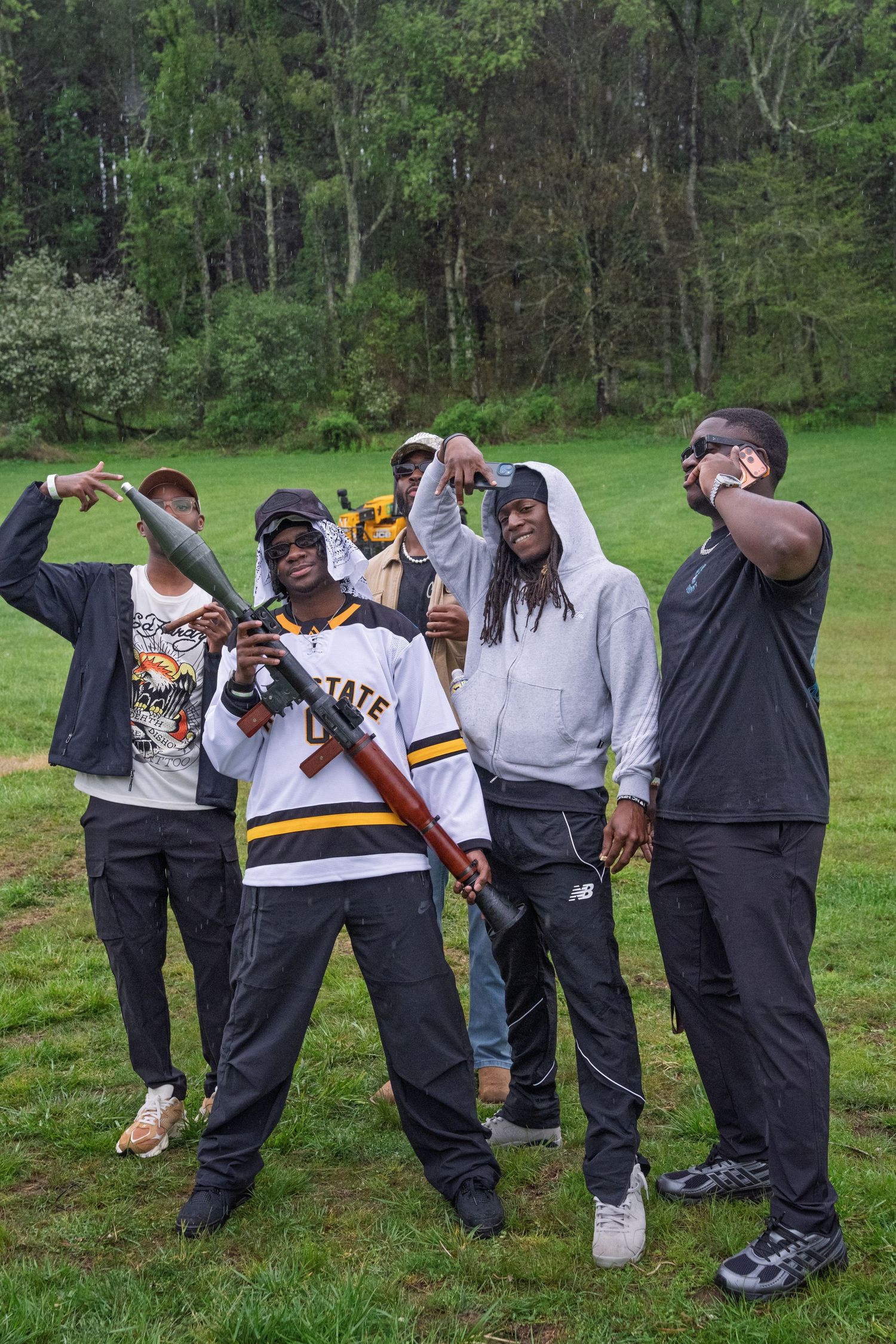 A group of five men pose together outdoors, one holding a prop rifle at a casual gathering.