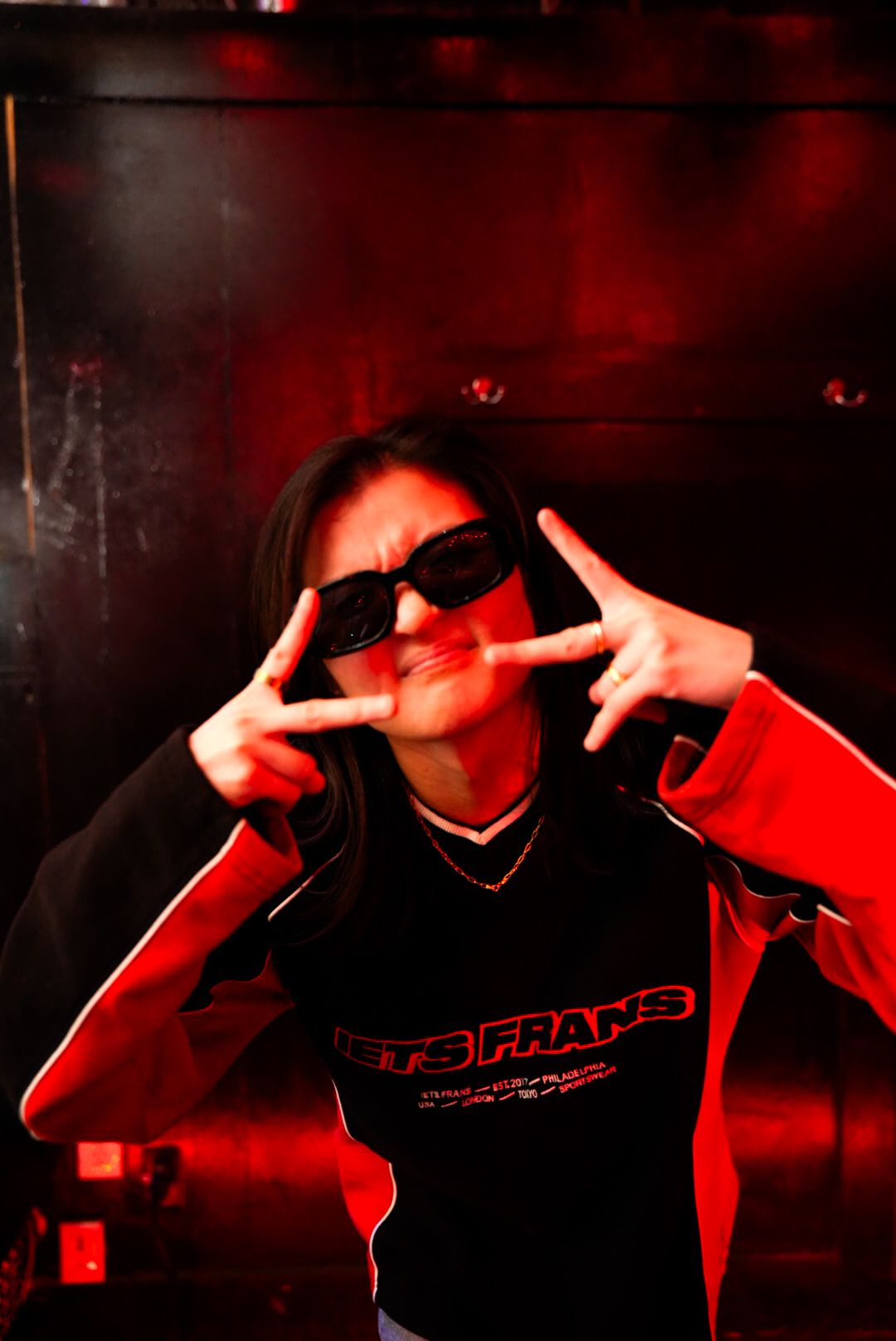 Person in Jets Frans jacket throwing peace signs under red club lighting.