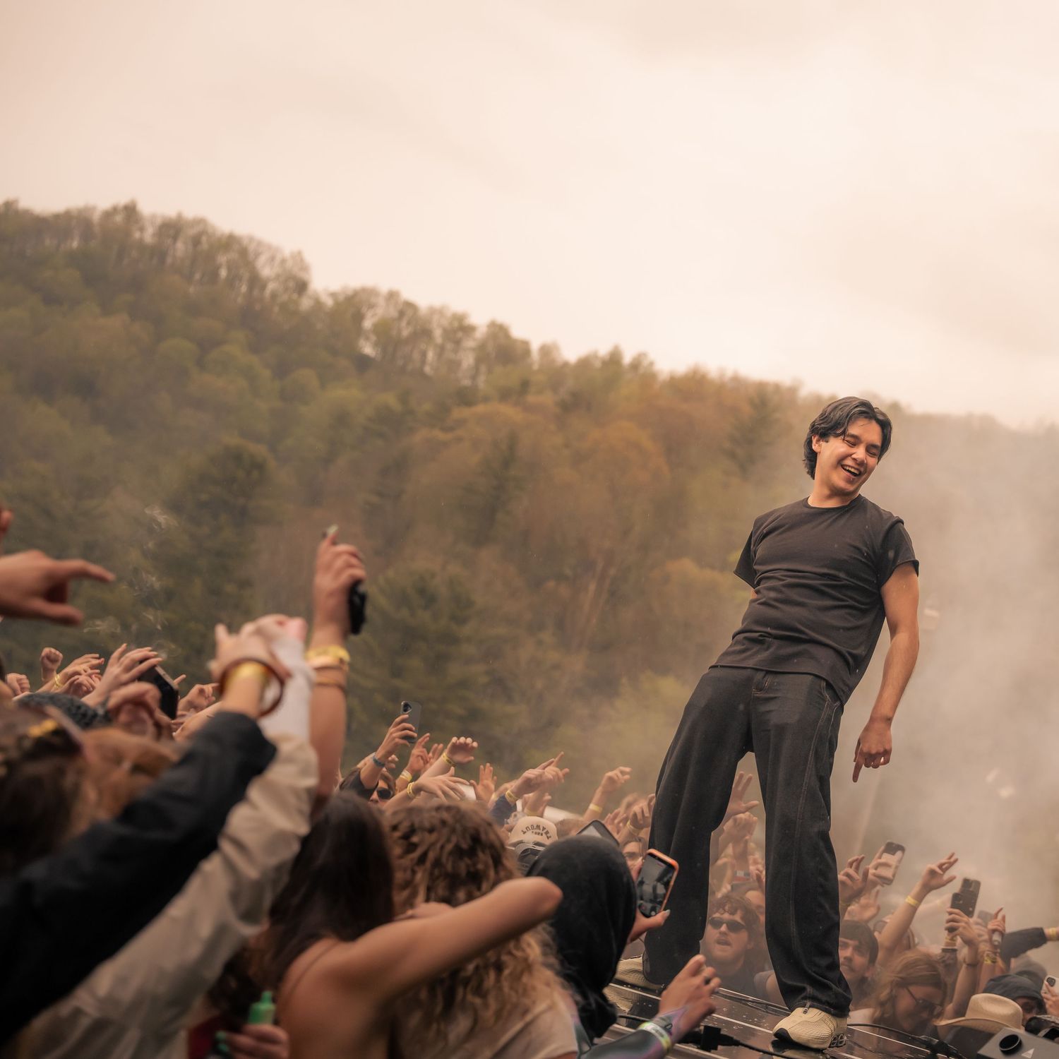 Performer standing elevated above crowd with arms out at a smoky outdoor music festival.