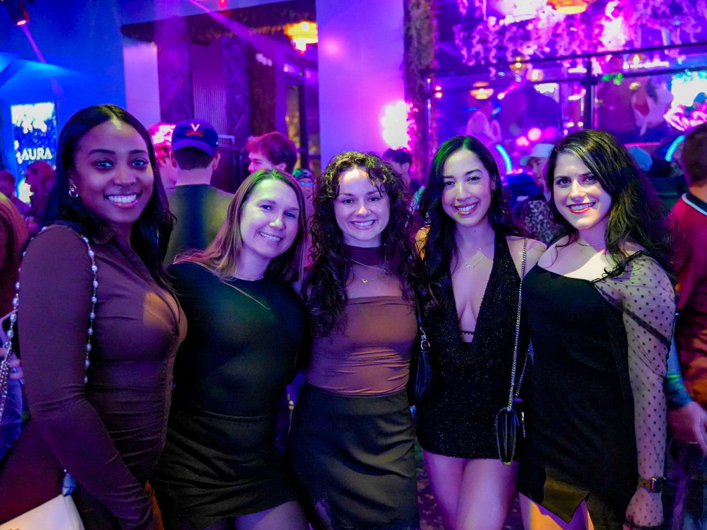 Five women pose together smiling at a vibrant nightclub with colorful purple and pink lighting.