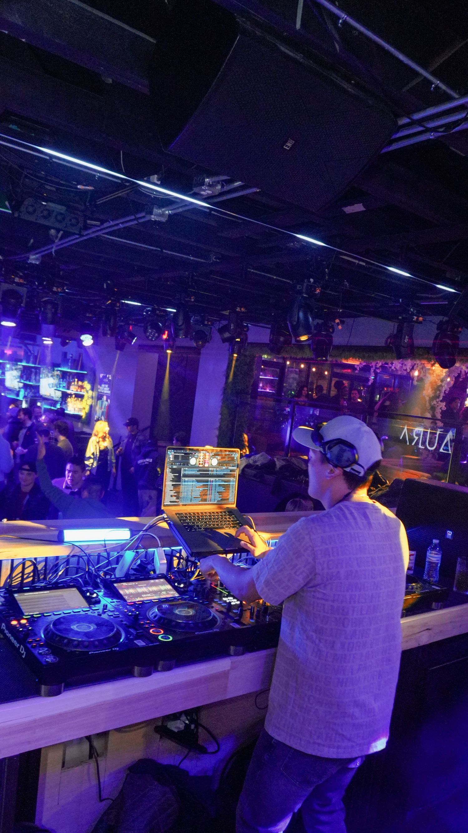 DJ performing at a nightclub booth with Pioneer decks under purple neon lights before a large crowd.