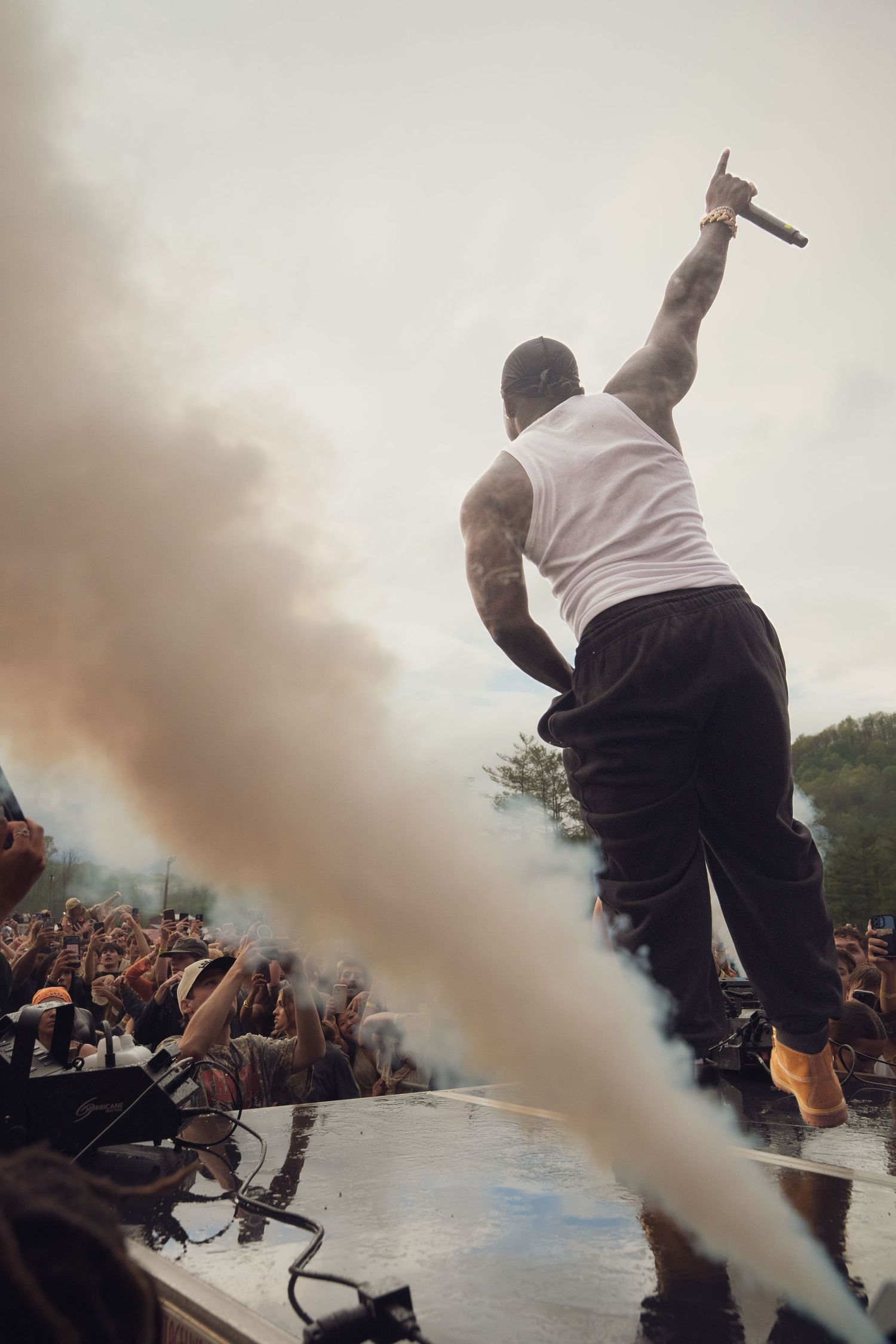 Muscular rapper on stage amid smoke effects, holding mic high toward massive outdoor concert crowd during festival performance.