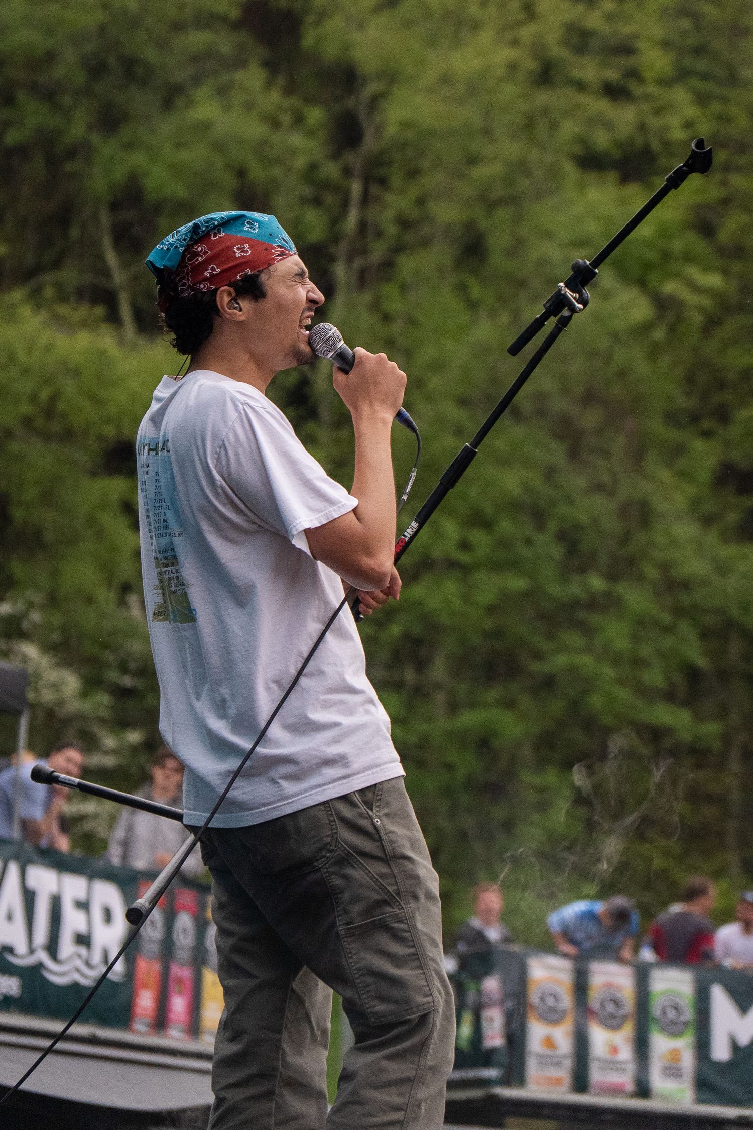 Young male performer singing into microphone on outdoor festival stage wearing white tee and red bandana.
