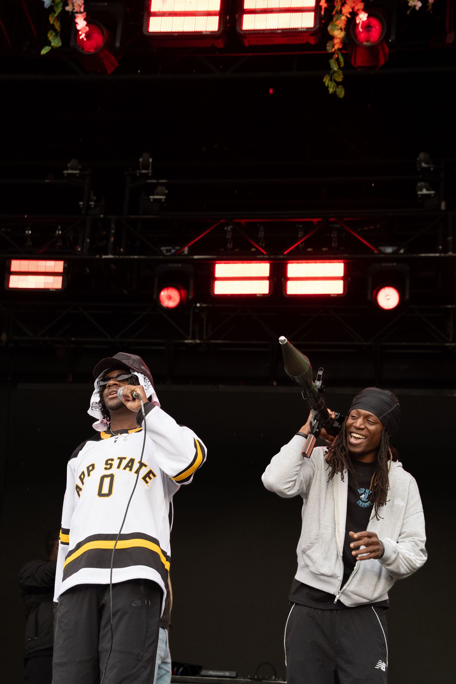 Two male hip-hop artists performing on outdoor festival stage, one in App State jersey.