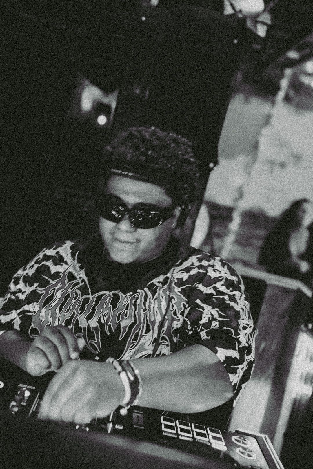 Black and white close-up of a DJ wearing sunglasses operating equipment at a nightclub event.