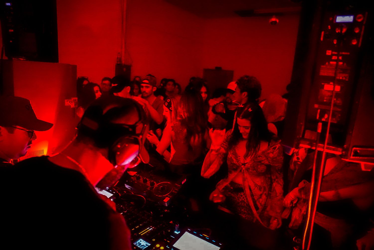 Energetic crowd dancing at a red-lit underground club party with DJ equipment in foreground.