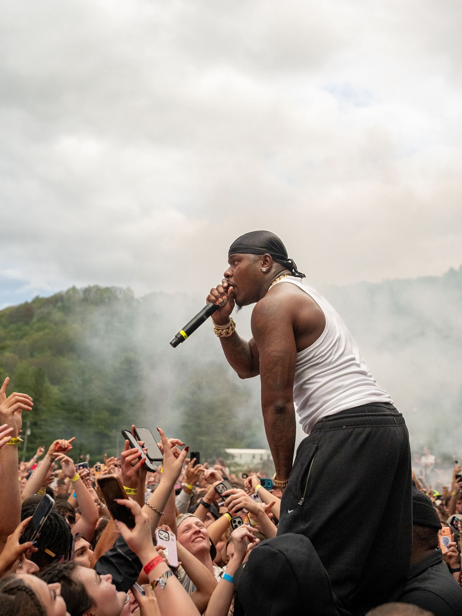 Male hip-hop artist in white tank top sings into microphone while crowd-surfing at smoky outdoor festival.