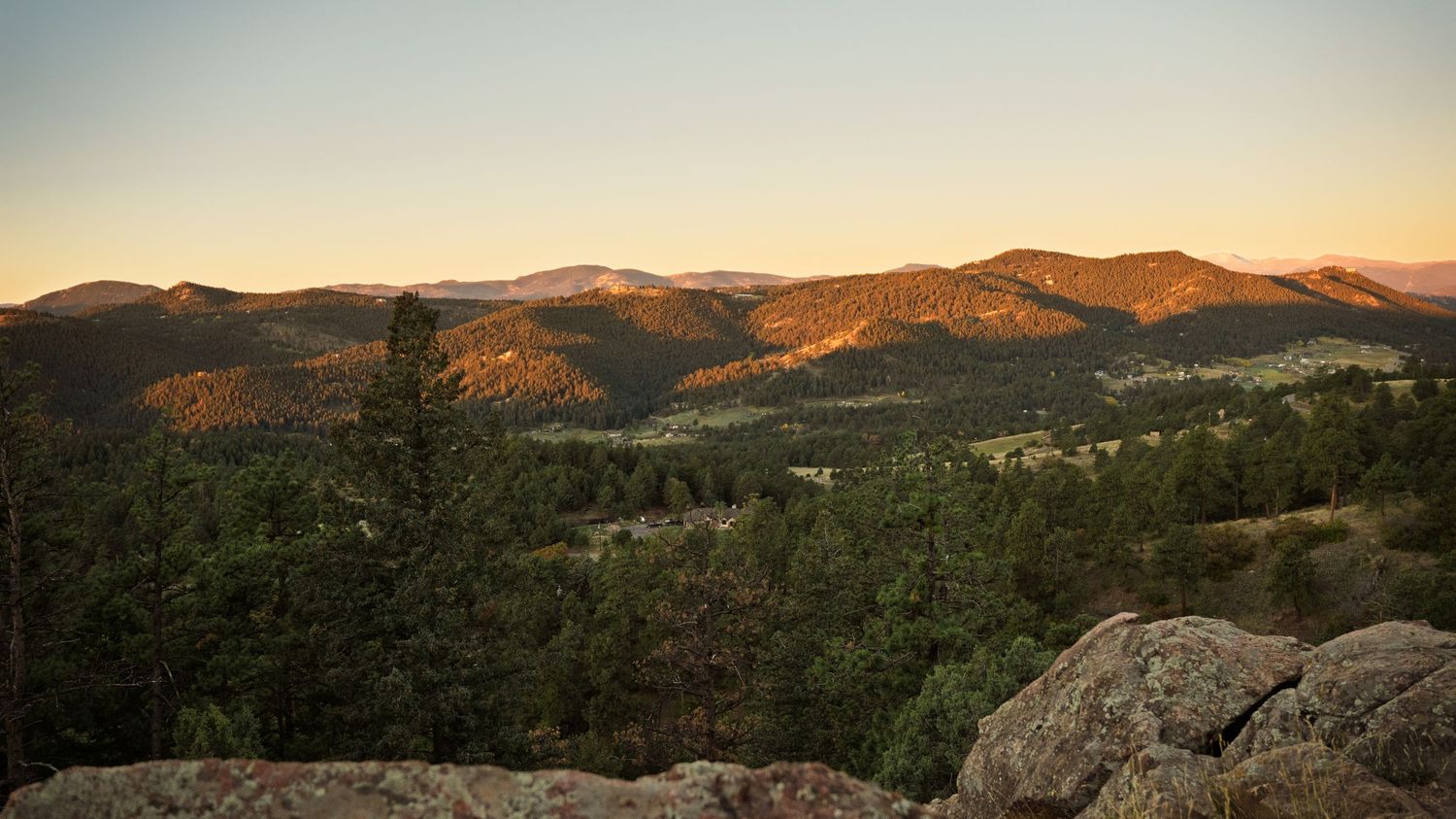 Golden sunset over pine tree covered mountain range with rocky foreground landscape view.
