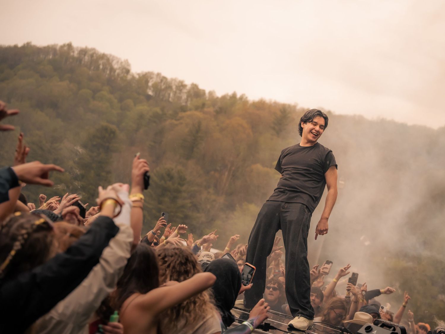 A young performer stands atop a platform crowd surfing at a smoky outdoor music festival.