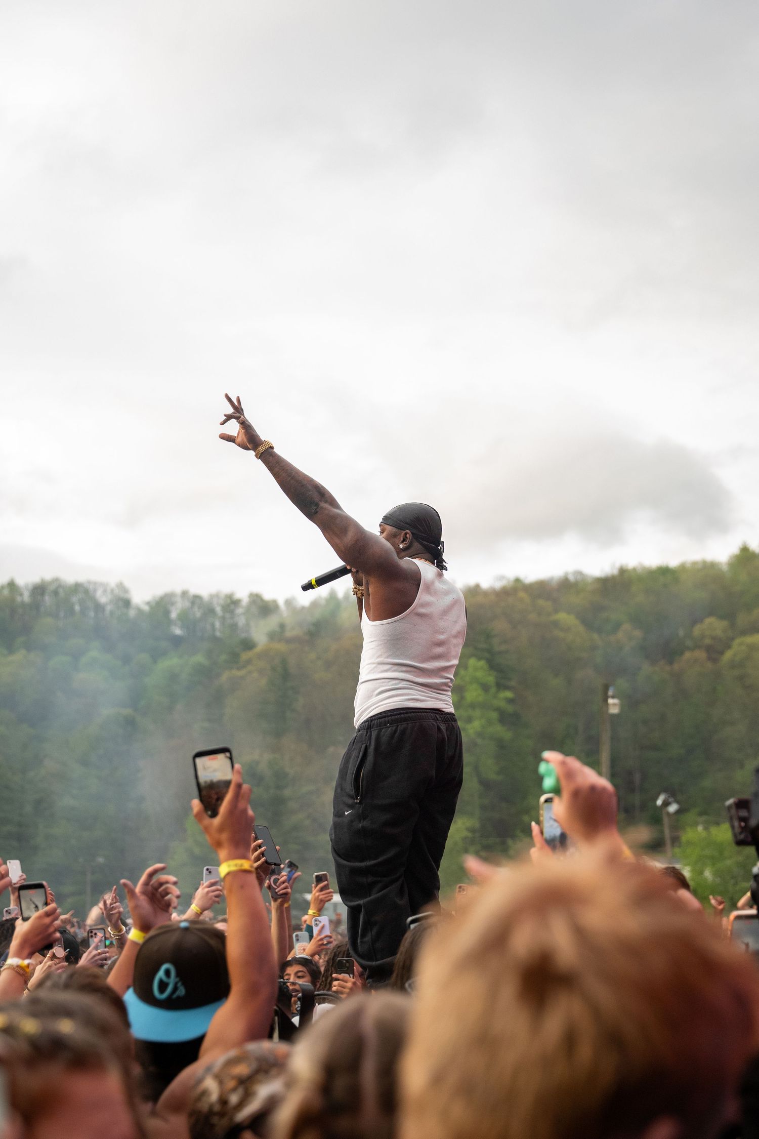Rapper raises hand with peace sign above energetic crowd at outdoor music festival.