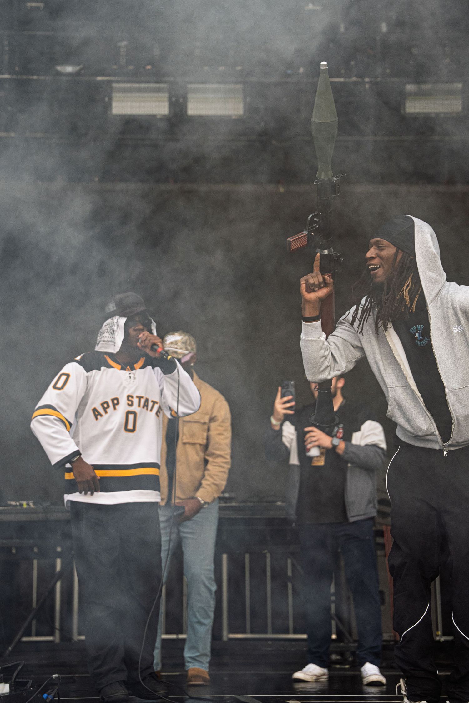 Two rappers perform on smoky outdoor stage, one in App State jersey, crowd visible behind.