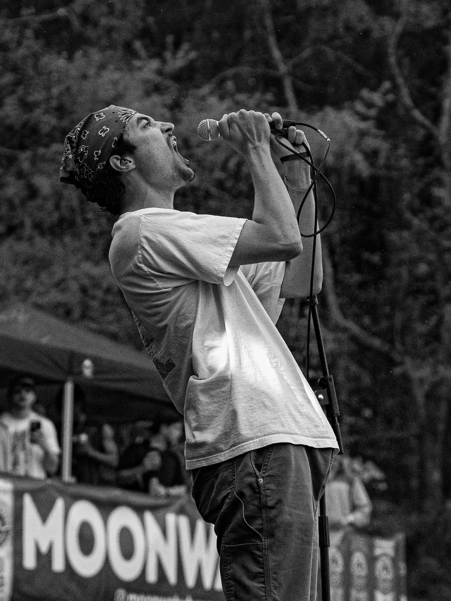 Black and white photo of male singer belting into mic at outdoor festival stage.