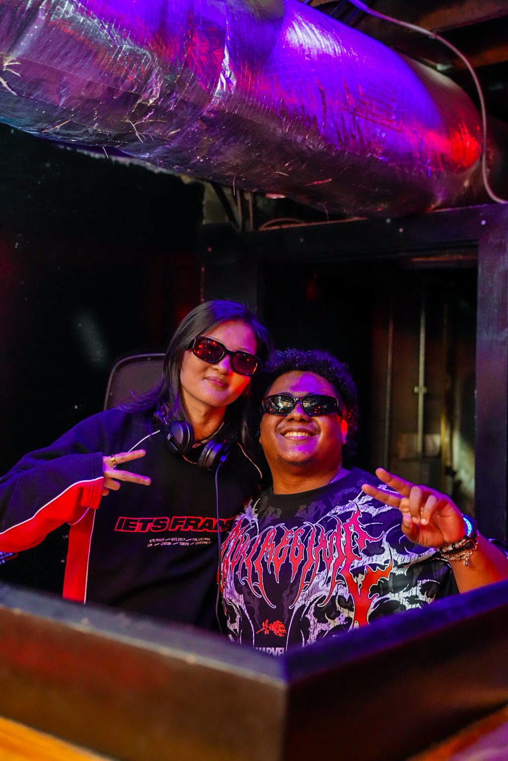 Two DJs wearing sunglasses pose together behind DJ equipment under purple neon lights at a nightclub.