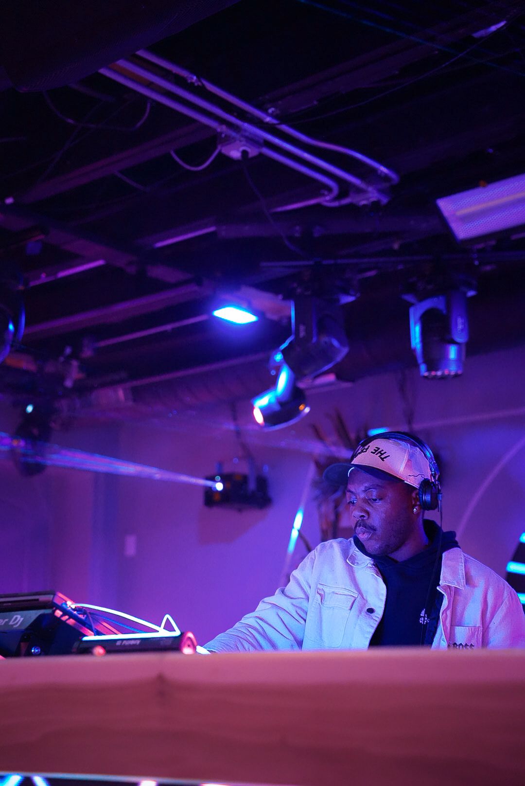 DJ in a white jacket and cap performing at a nightclub booth under vibrant purple stage lighting.