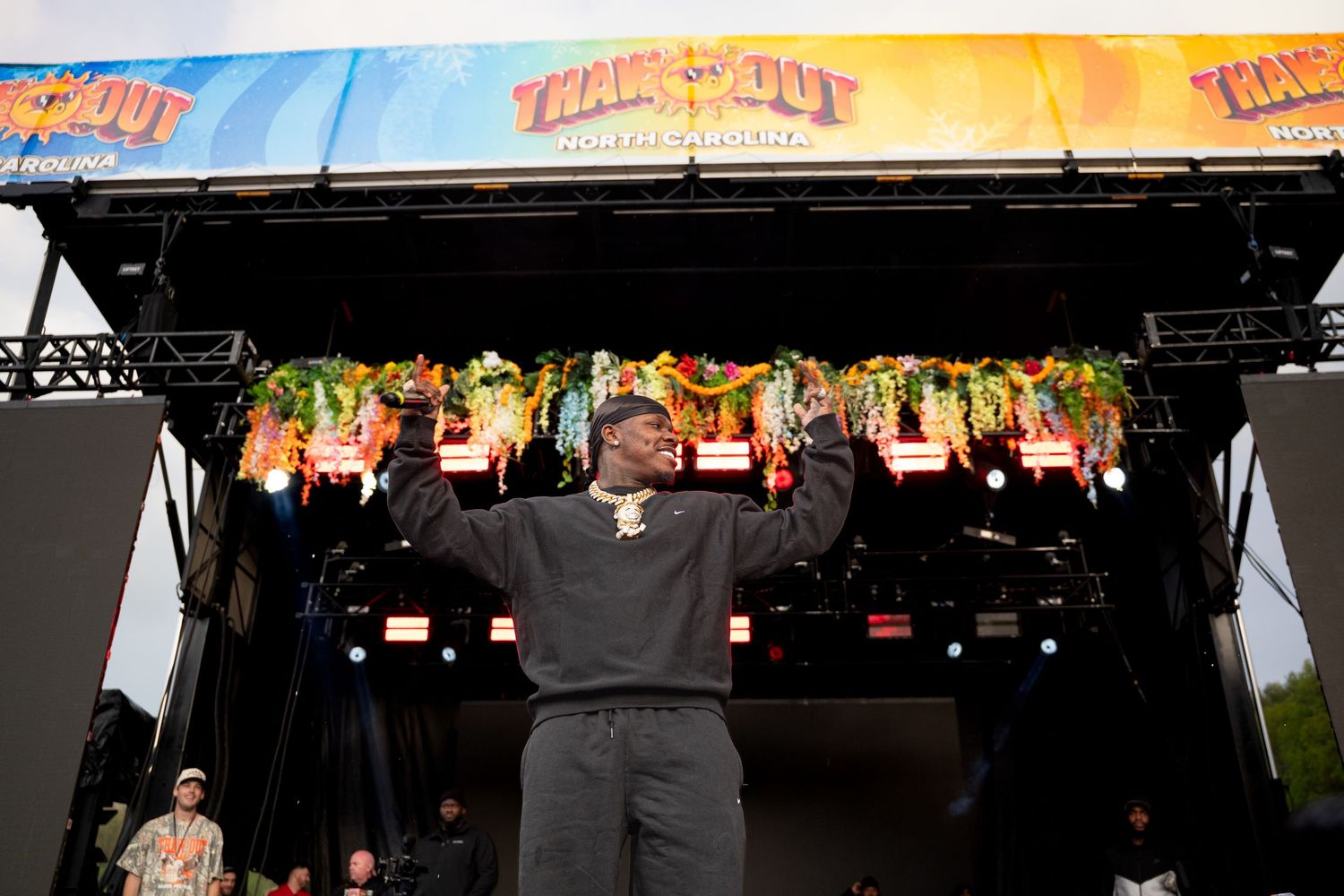 Rapper stands triumphantly on outdoor festival stage with arms raised before colorful floral-decorated TrackOut Music Festival backdrop.